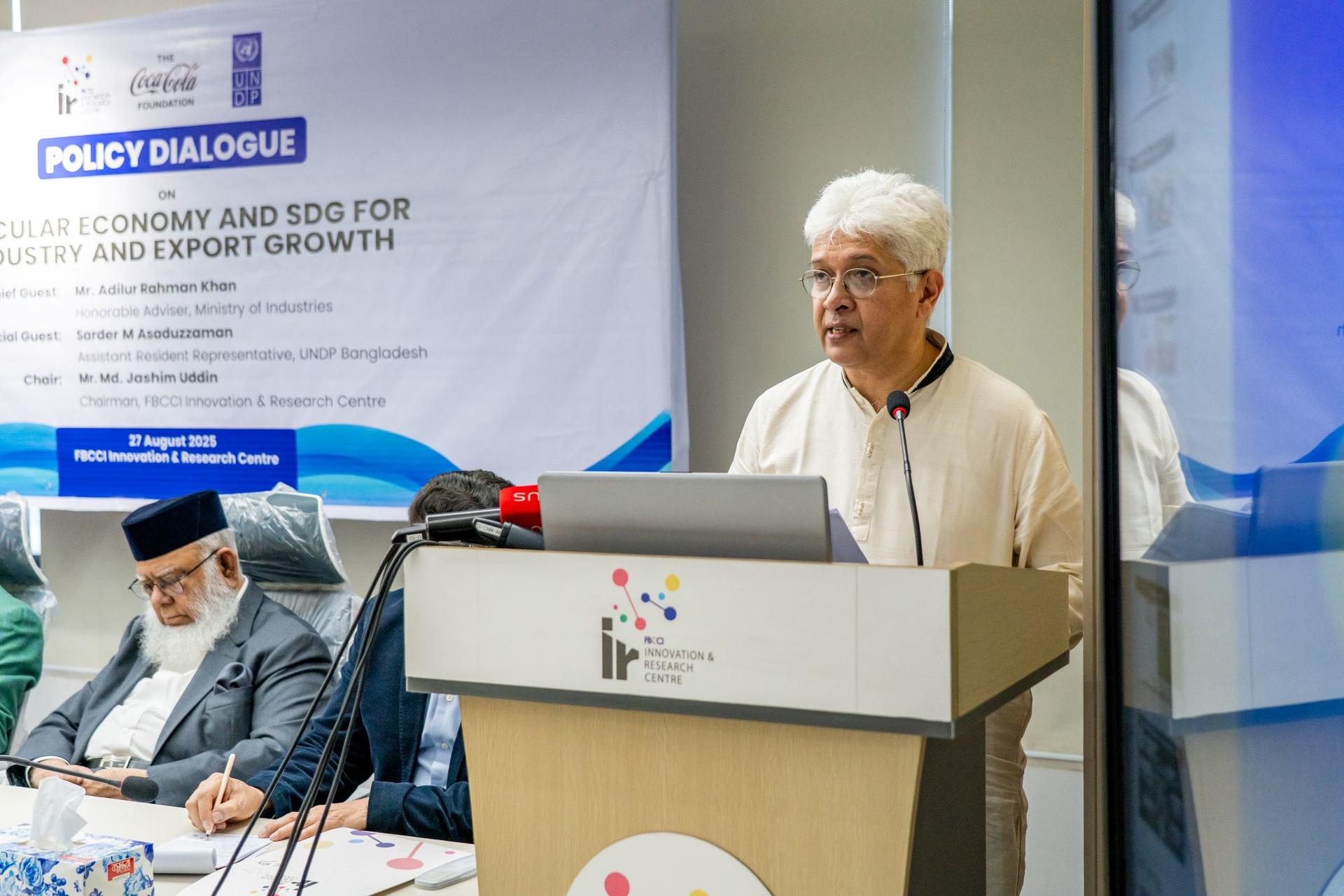 A speaker delivers a presentation at a policy dialogue event, with attendees seated nearby.