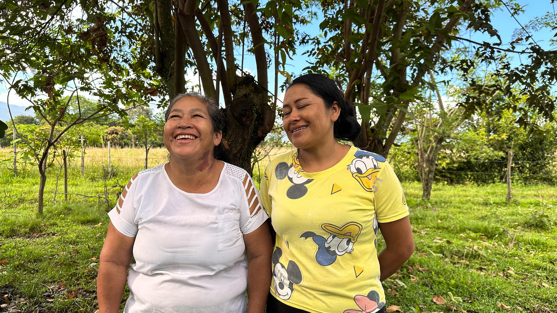 Two women smiling and laughing under a tree in a green outdoor setting.