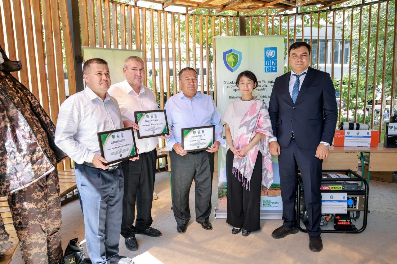 Five individuals stand together, holding certificates in a brightly lit setting with greenery.