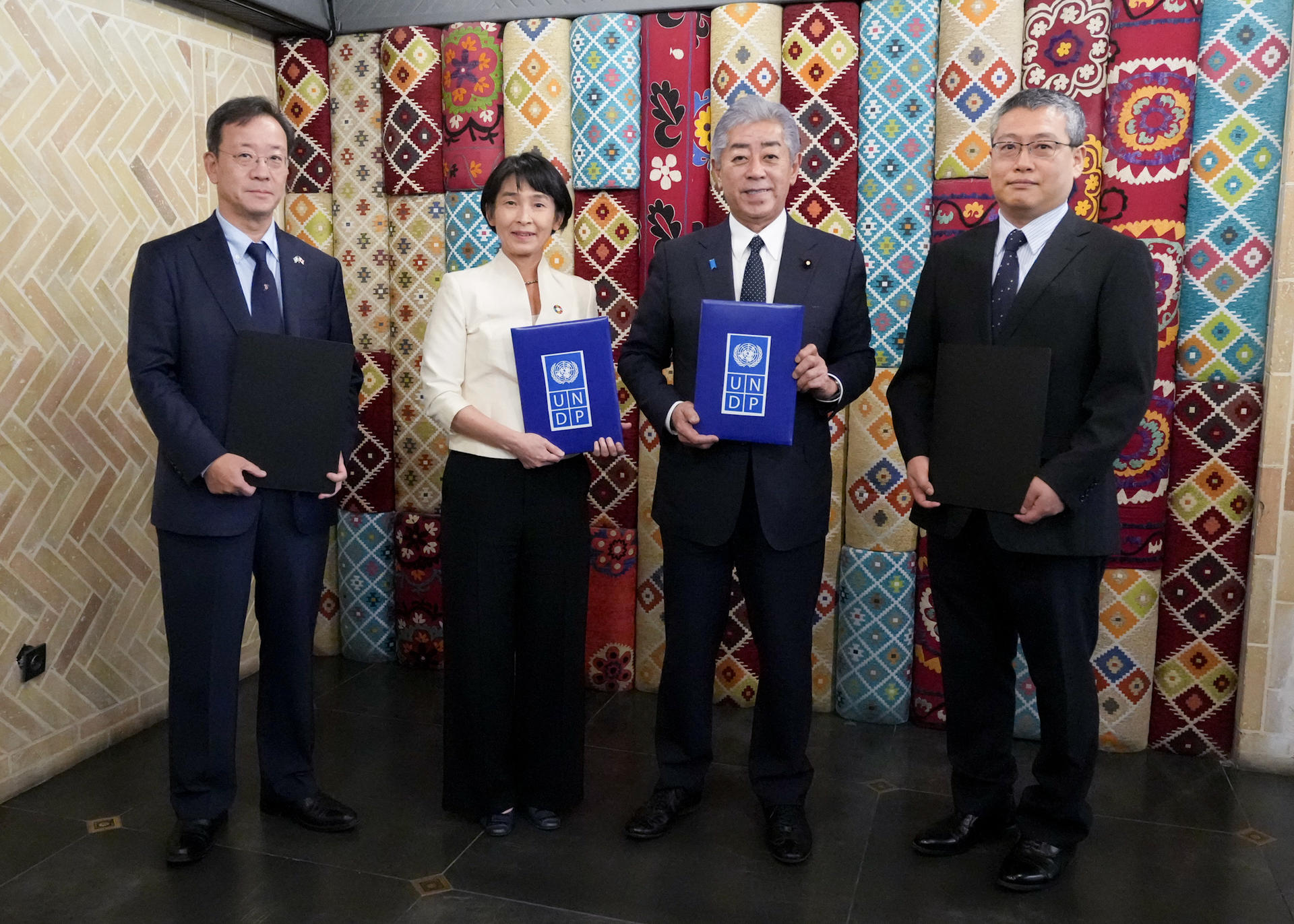 Four professionals stand together, holding blue documents, against a colorful patterned backdrop.