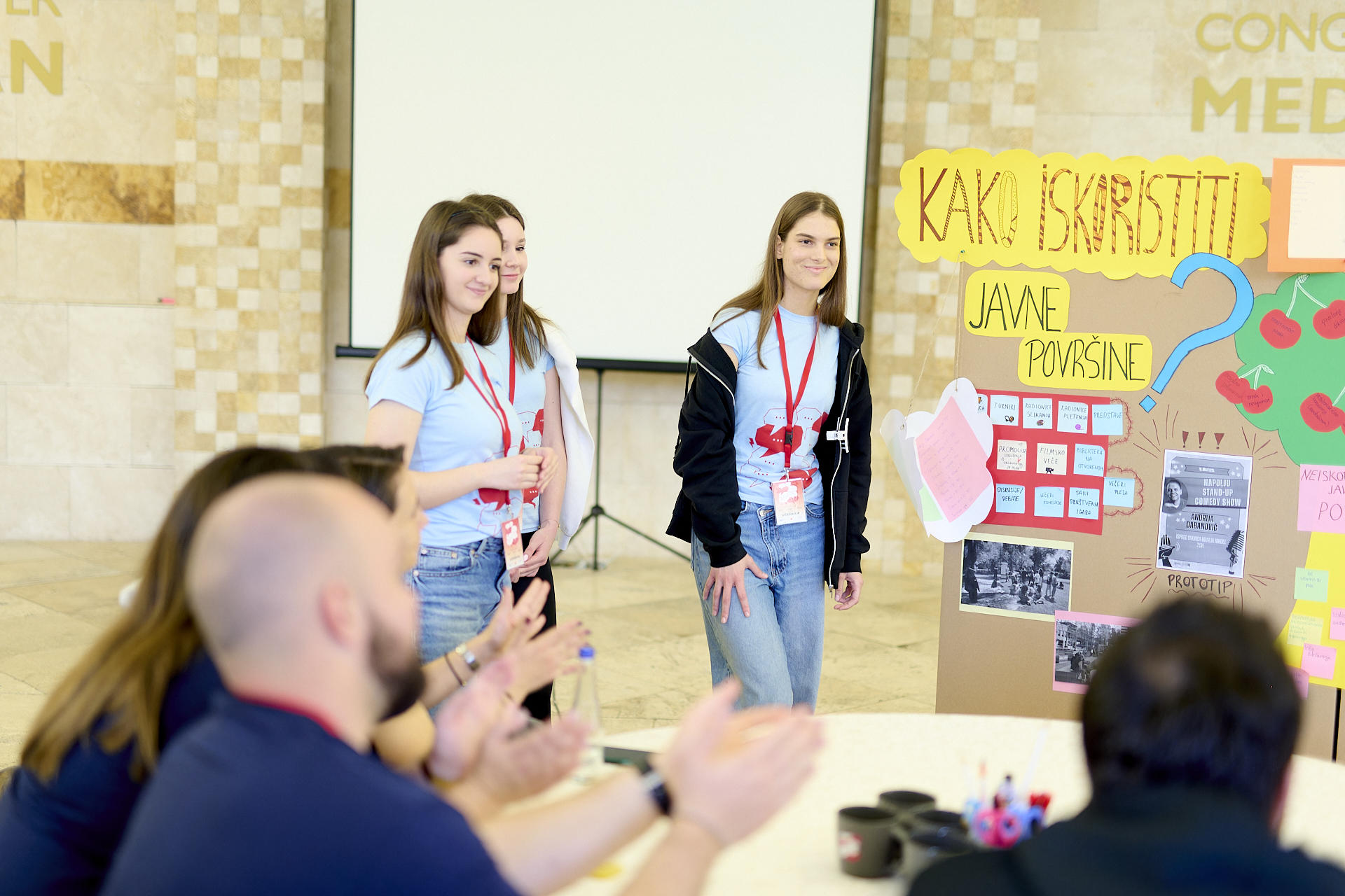 Three young women presenting in front of an audience at a workshop with colorful posters.