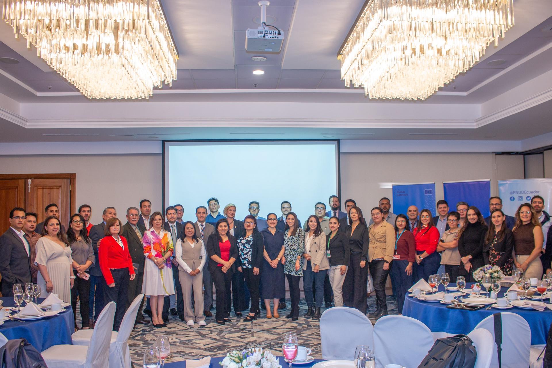 A group photo of diverse attendees in a conference room with chandeliers and a presentation screen.