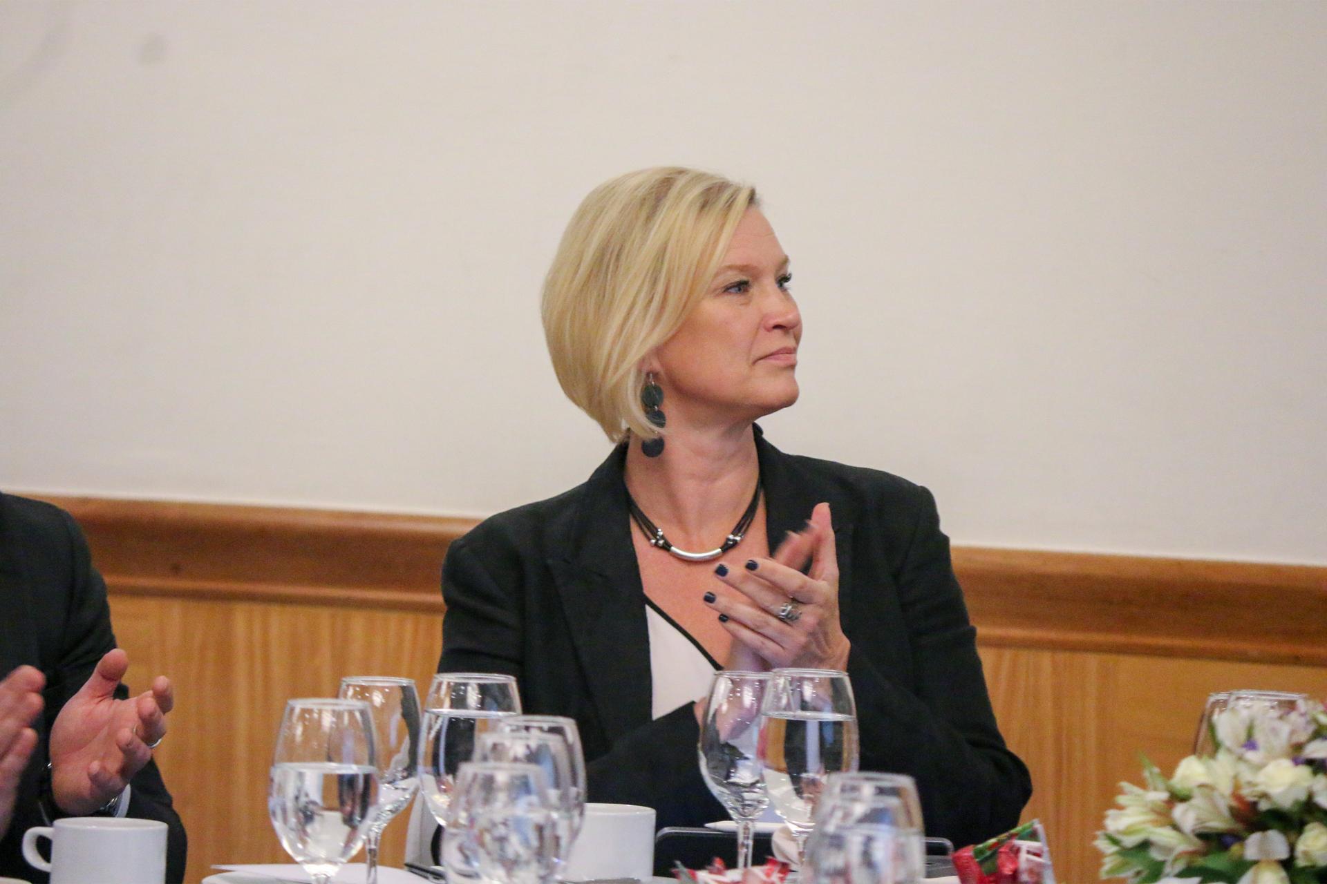 A woman with short blonde hair sits at a table, turning her head slightly, looking thoughtful.