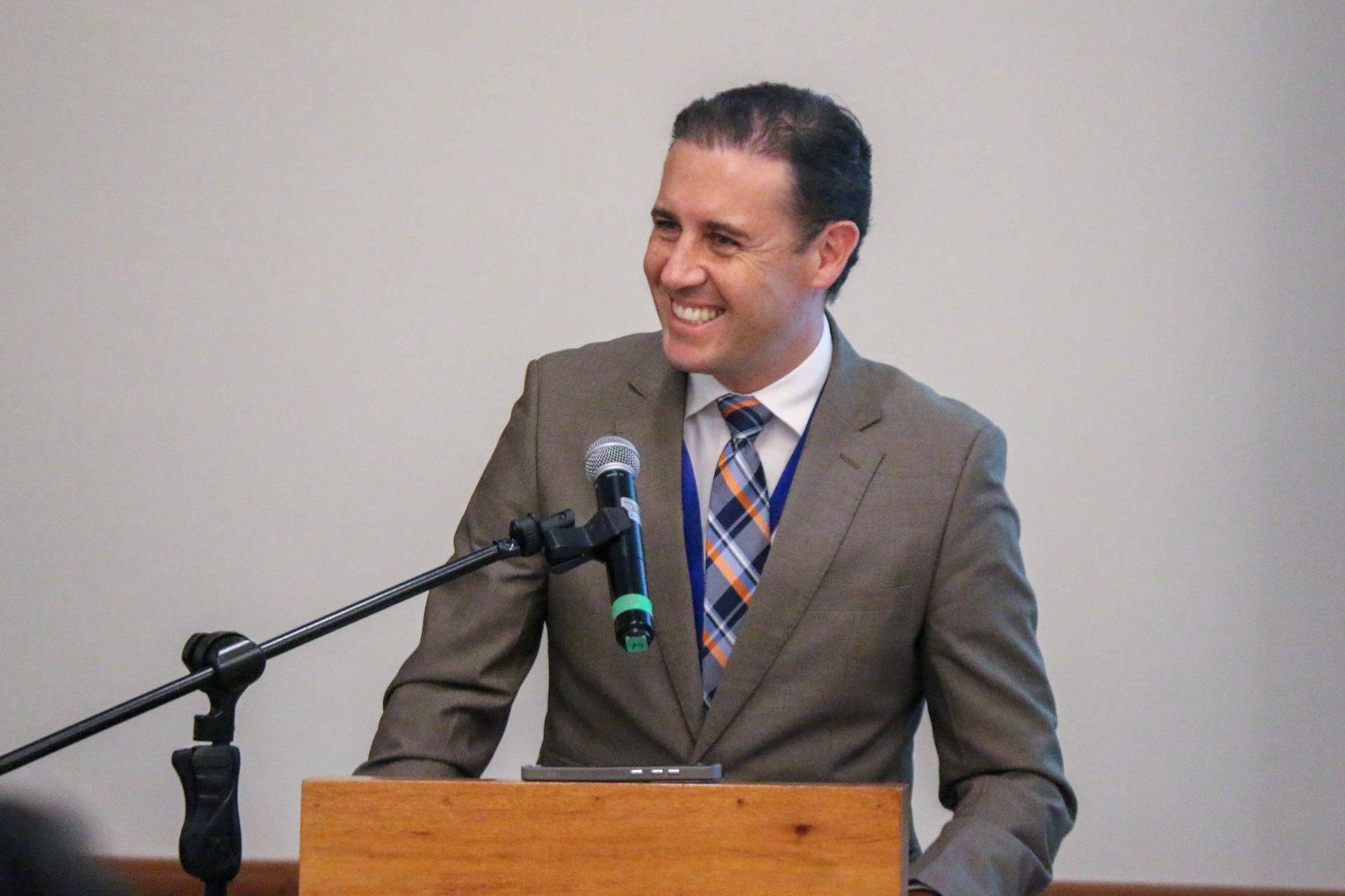 Smiling man in a suit stands at a podium with a microphone, looking engaged.
