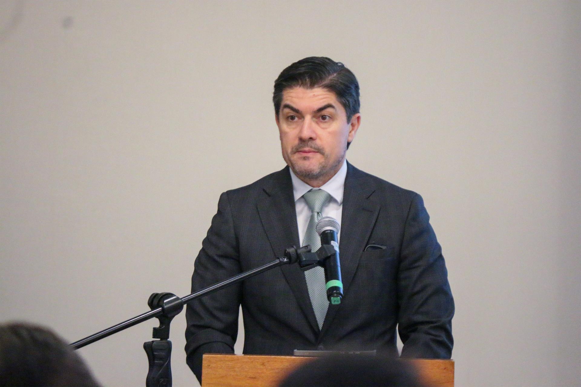 Man in a suit stands at a podium with a microphone, looking serious in a conference setting.