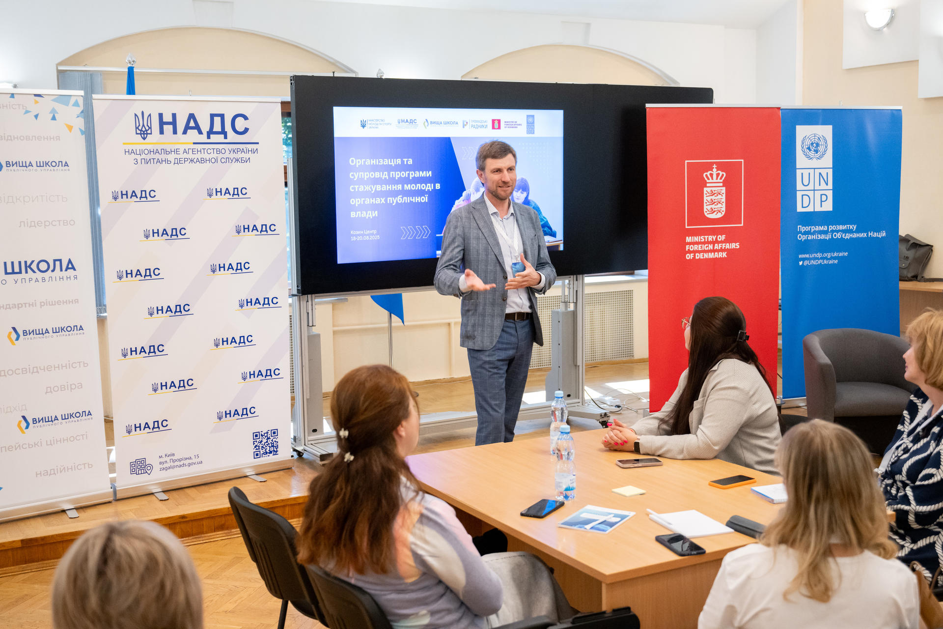 A speaker presents to an audience in a conference room, with colorful banners and a screen behind.