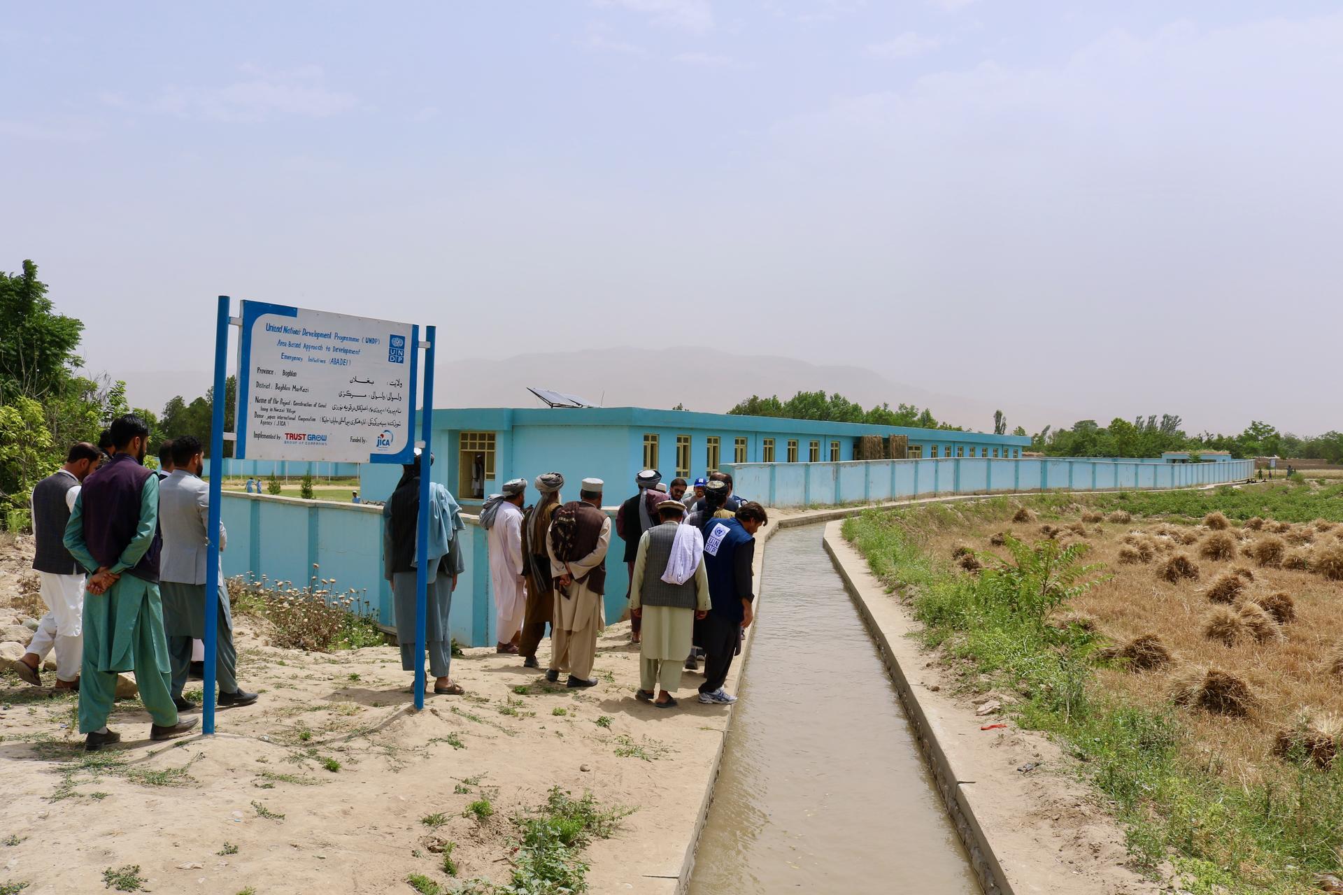 Group of people gathering near a blue information sign along a path by the water.