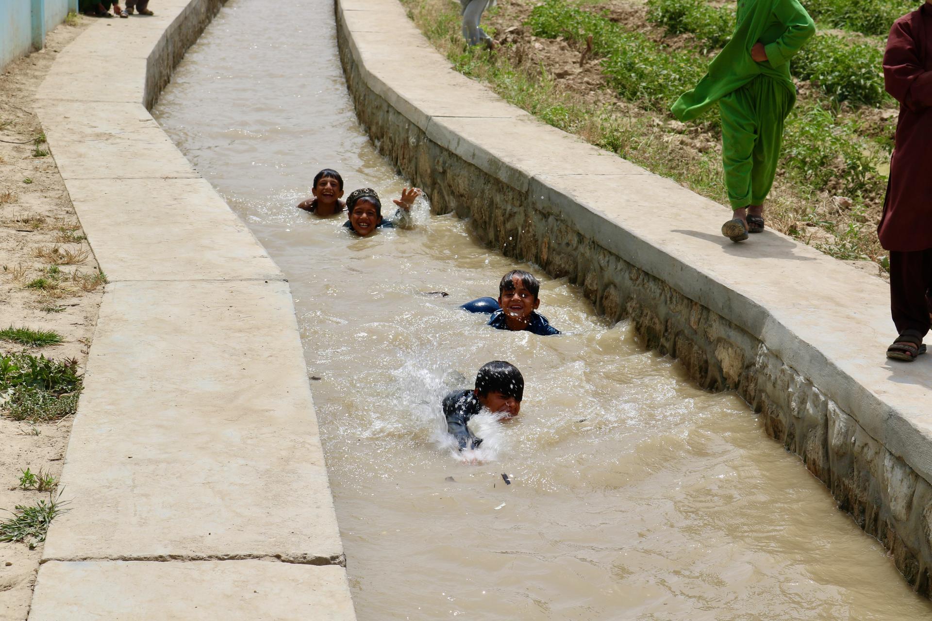 Children wading and playing in a sunlit, narrow concrete canal filled with muddy water.