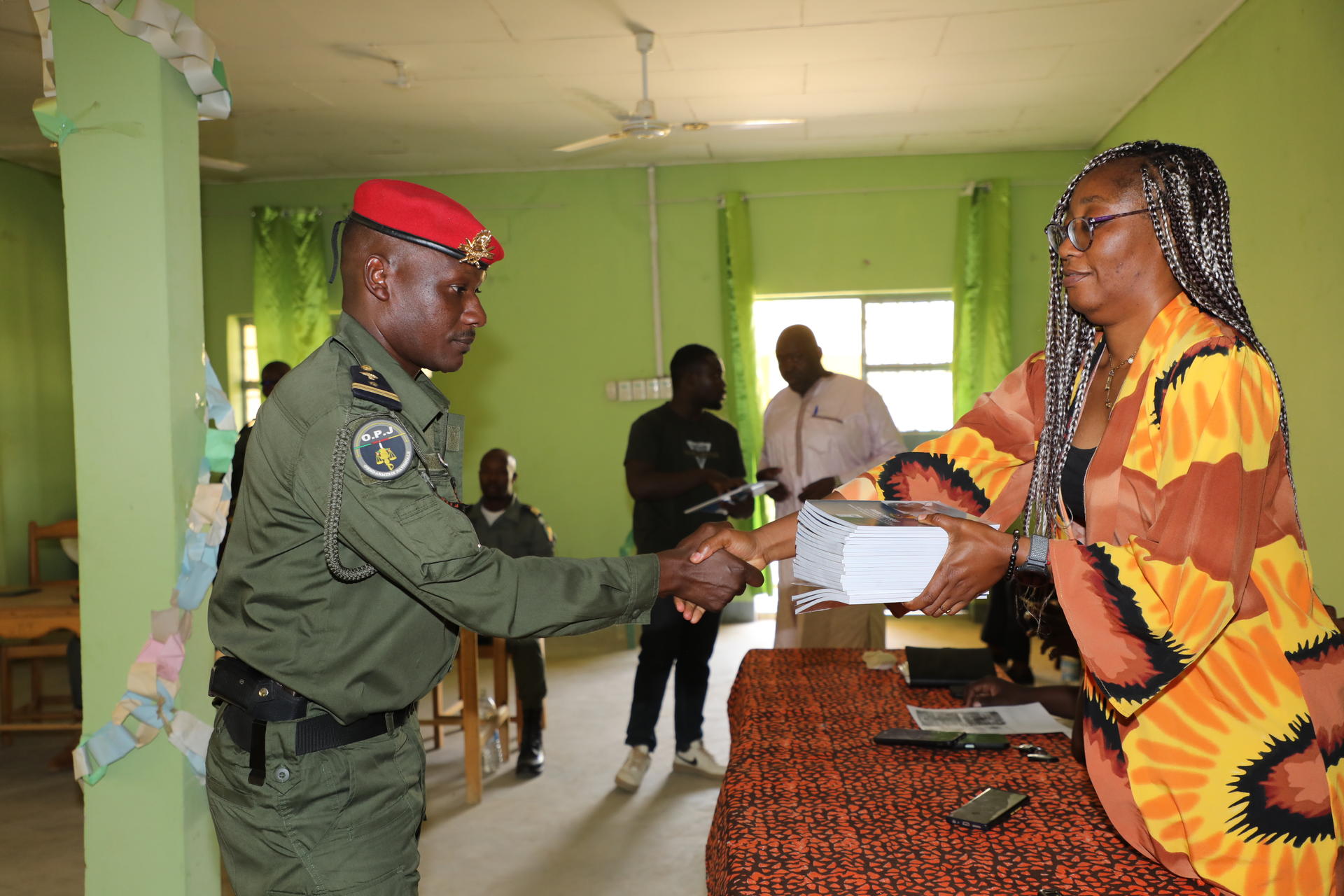 Photograph of a uniformed officer handing a document to a woman in a bright orange dress at a table.