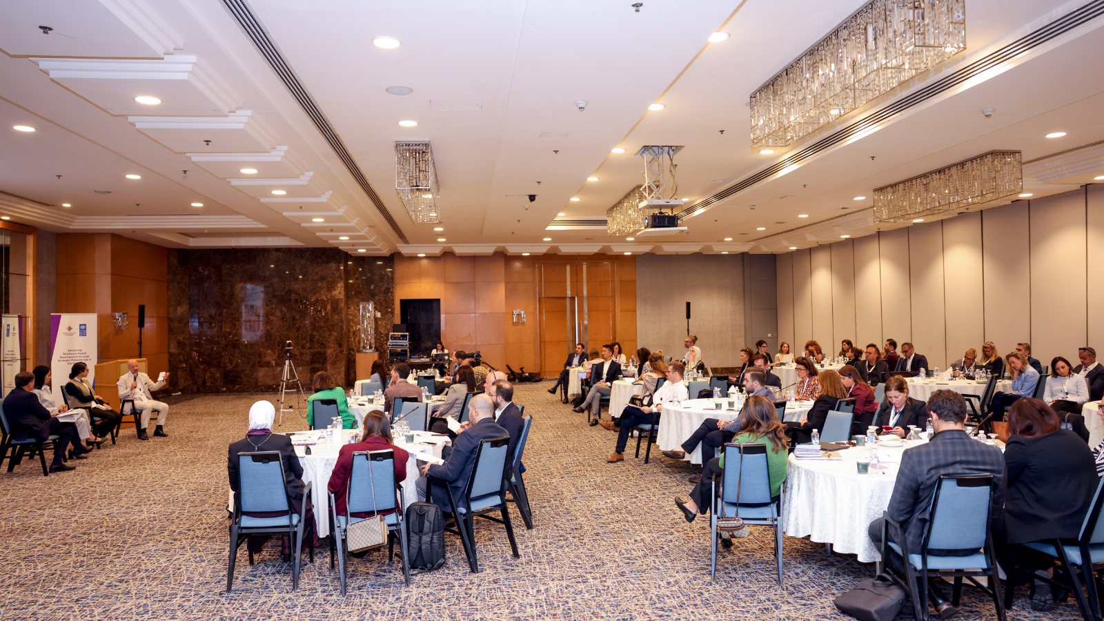 Well-lit banquet hall with round tables and attendees listening to a presentation.