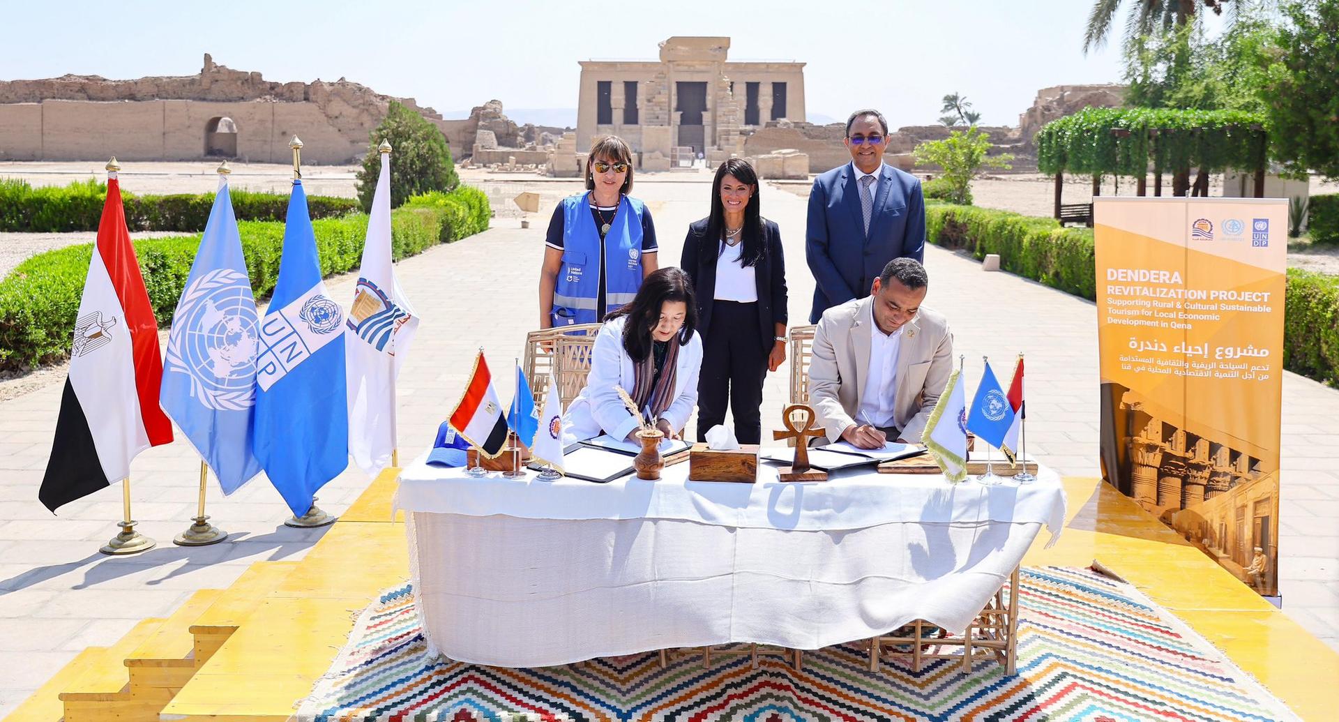 Group of people standing behind a table with a white cloth outdoors; flags and banners nearby.