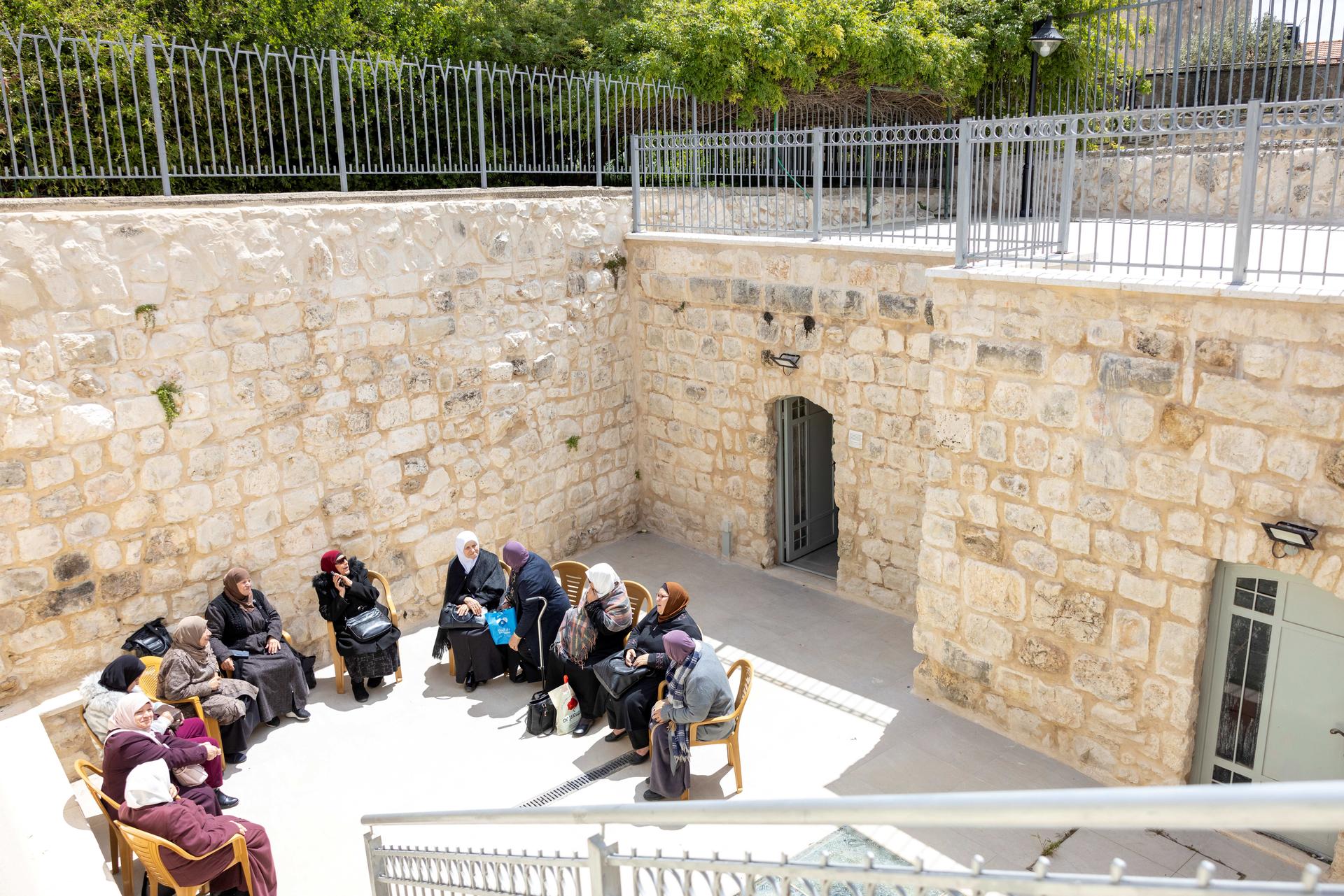 Group of people seated along a stone courtyard wall near an arched doorway.