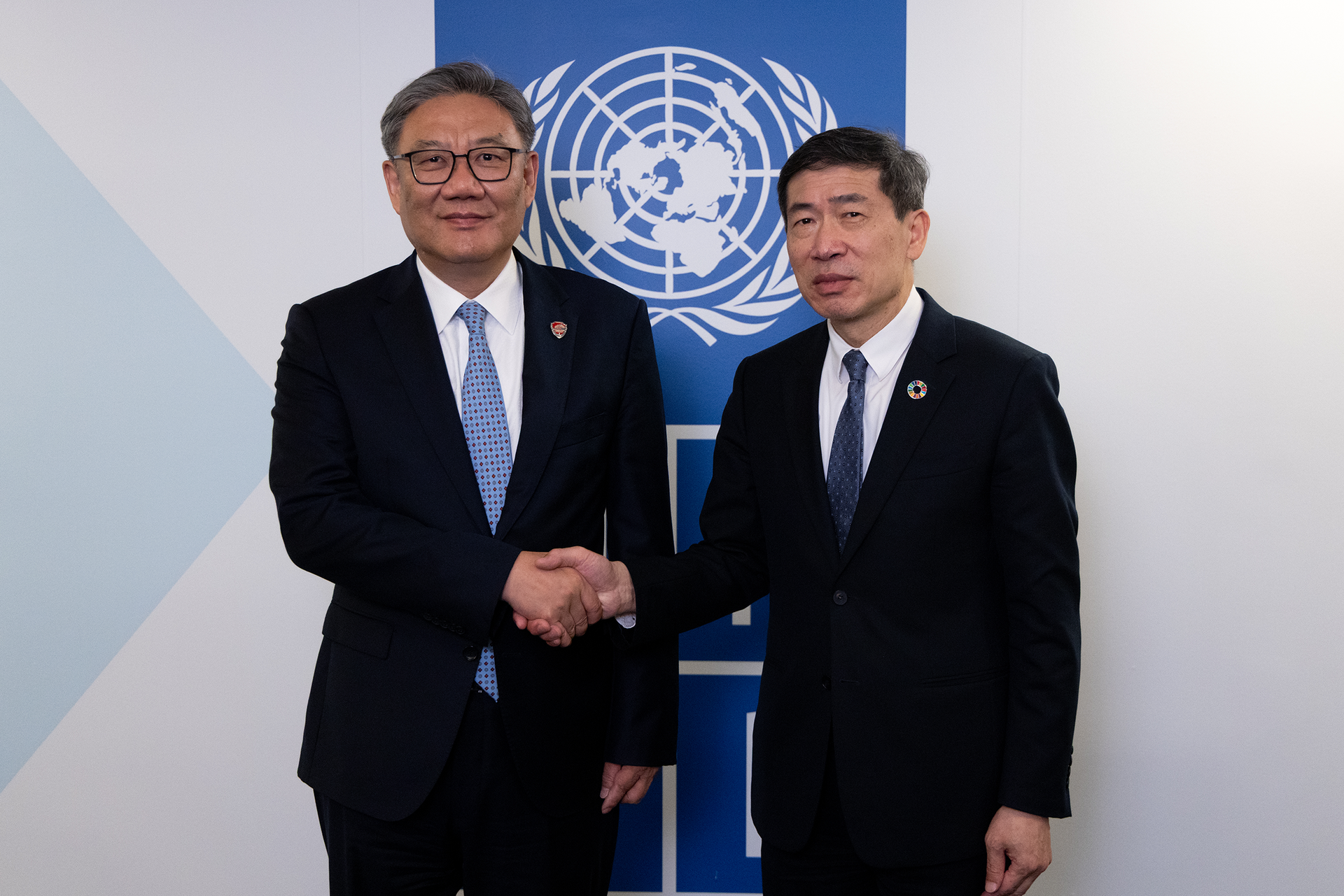 Two men in formal suits shake hands in front of a United Nations emblem backdrop.