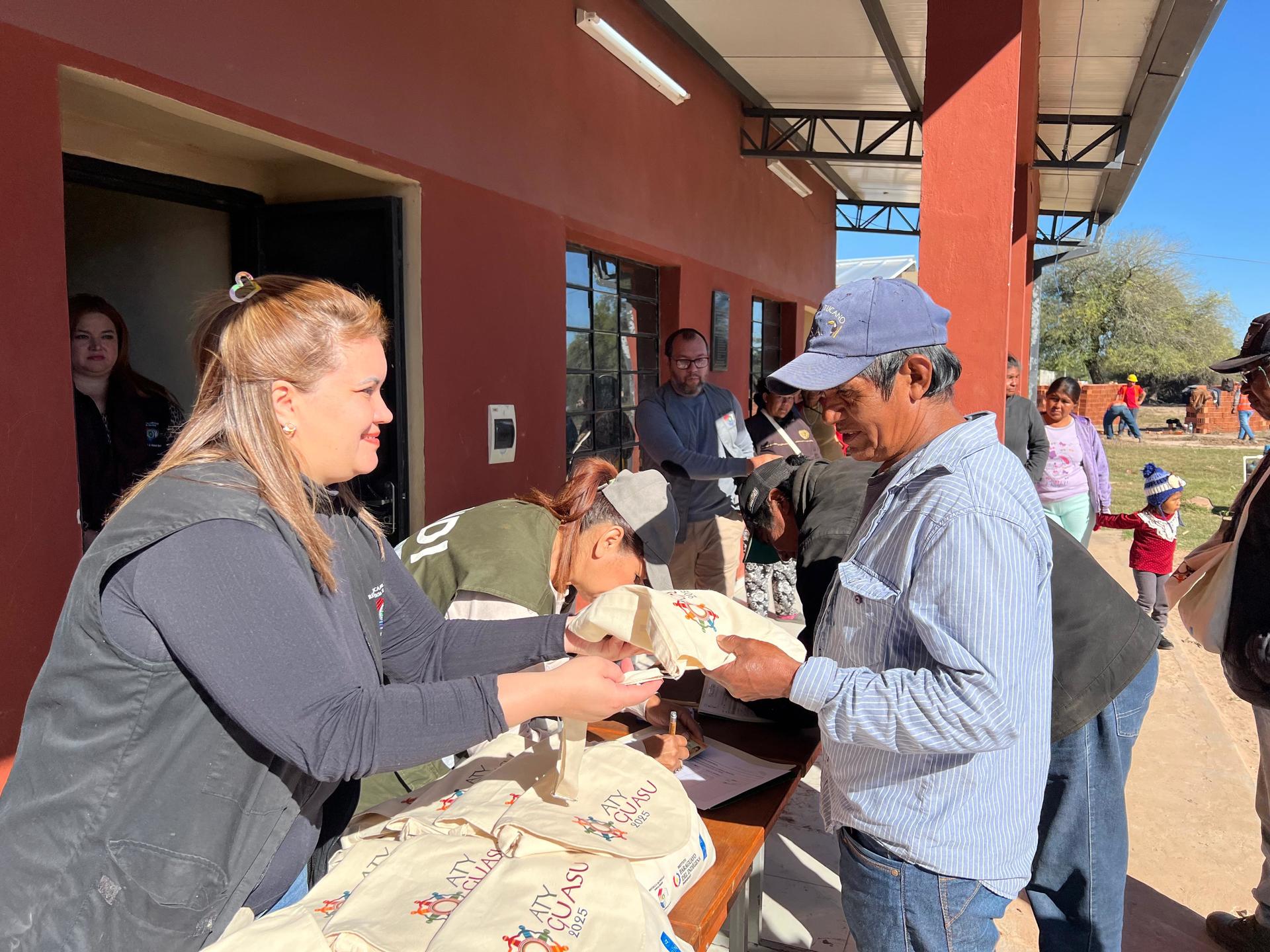 Volunteers hand out items at outdoor community event; table with patterned cloth near red building.
