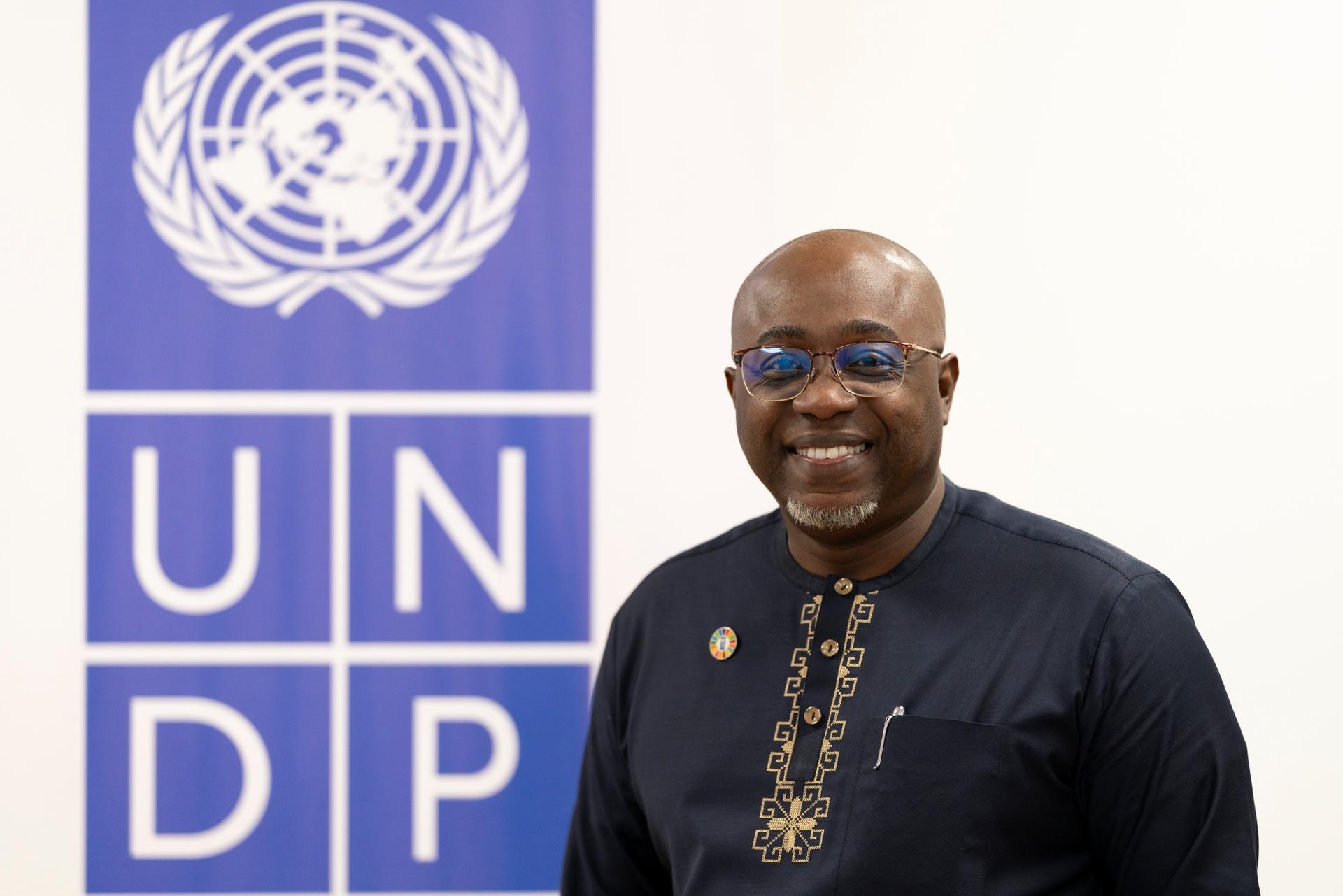 Man in dark tunic with gold embroidery stands beside a UNDP banner.