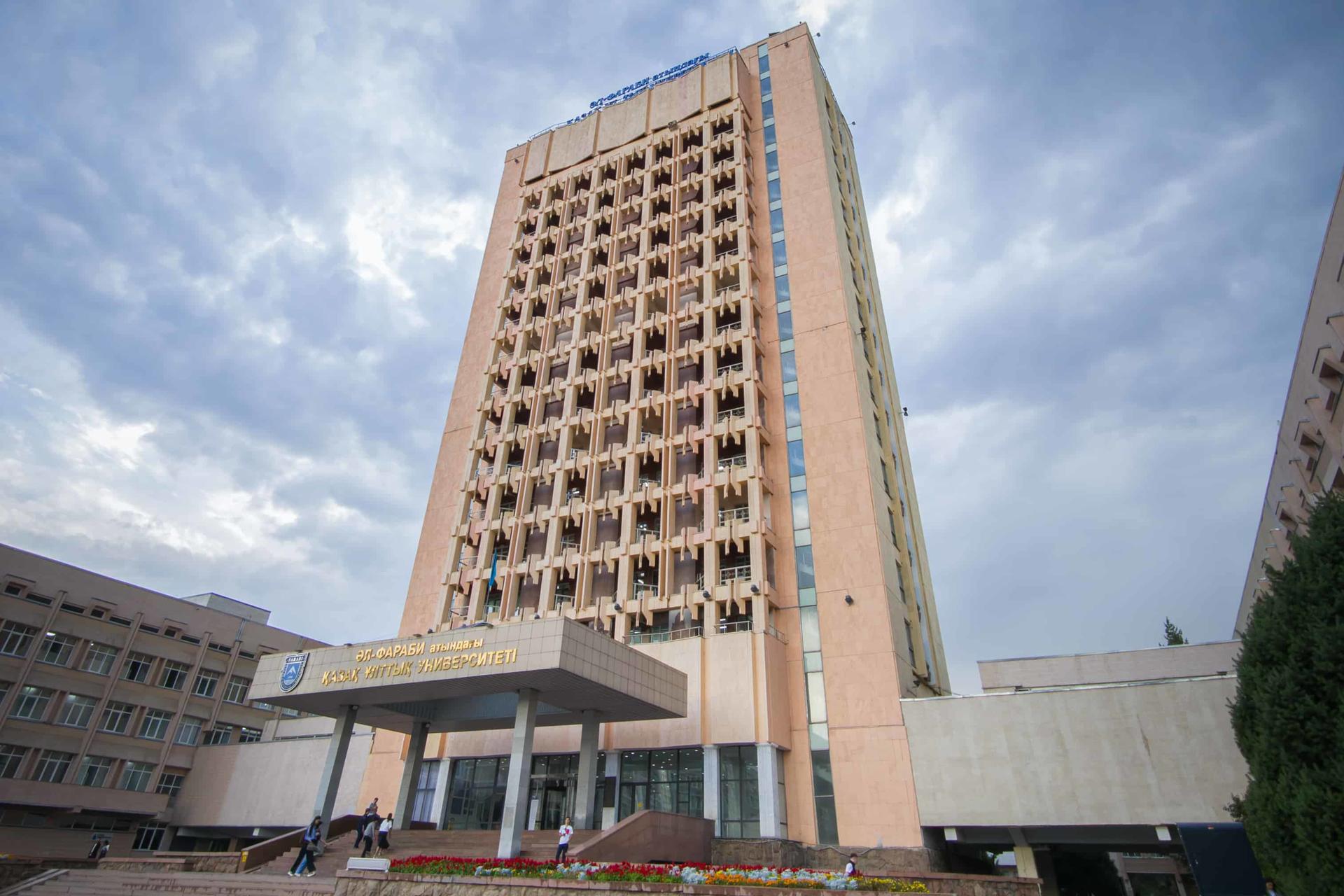 Tall beige high-rise with a glass canopy entrance, set among low buildings under a cloudy sky.