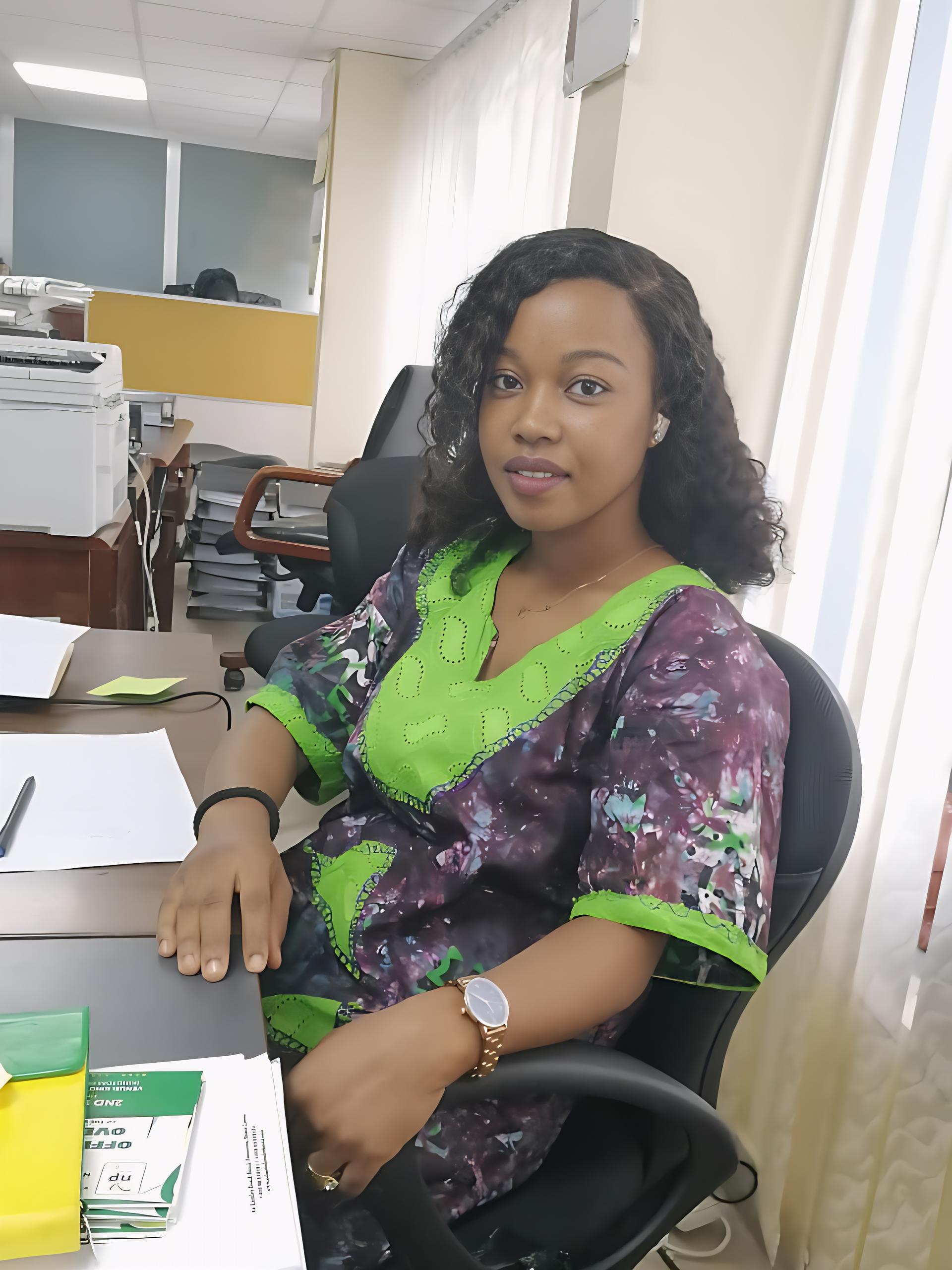 Woman seated at a desk in an office, papers nearby, wearing a purple-green patterned top.