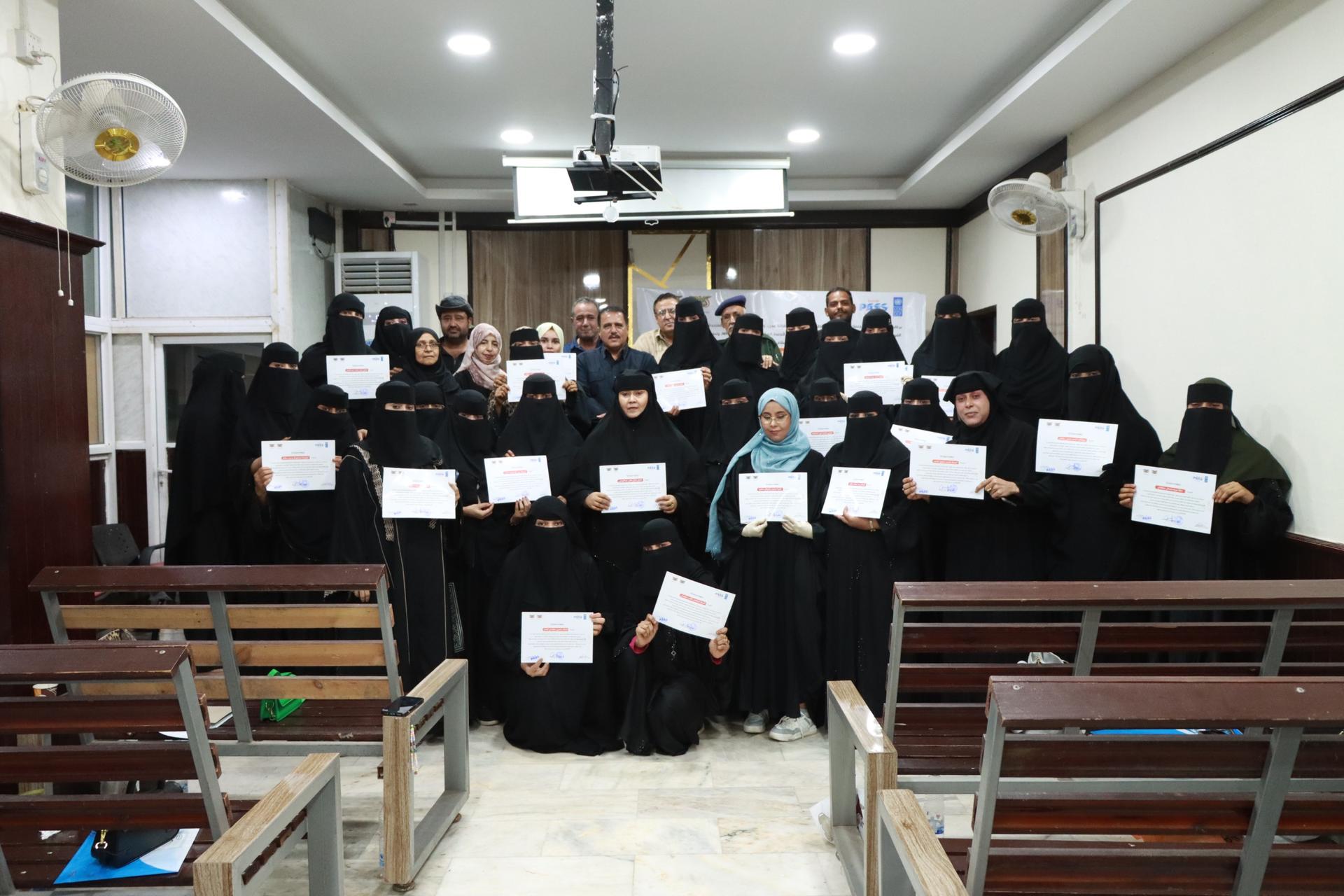 Group of graduates in black robes posing with certificates in a classroom.