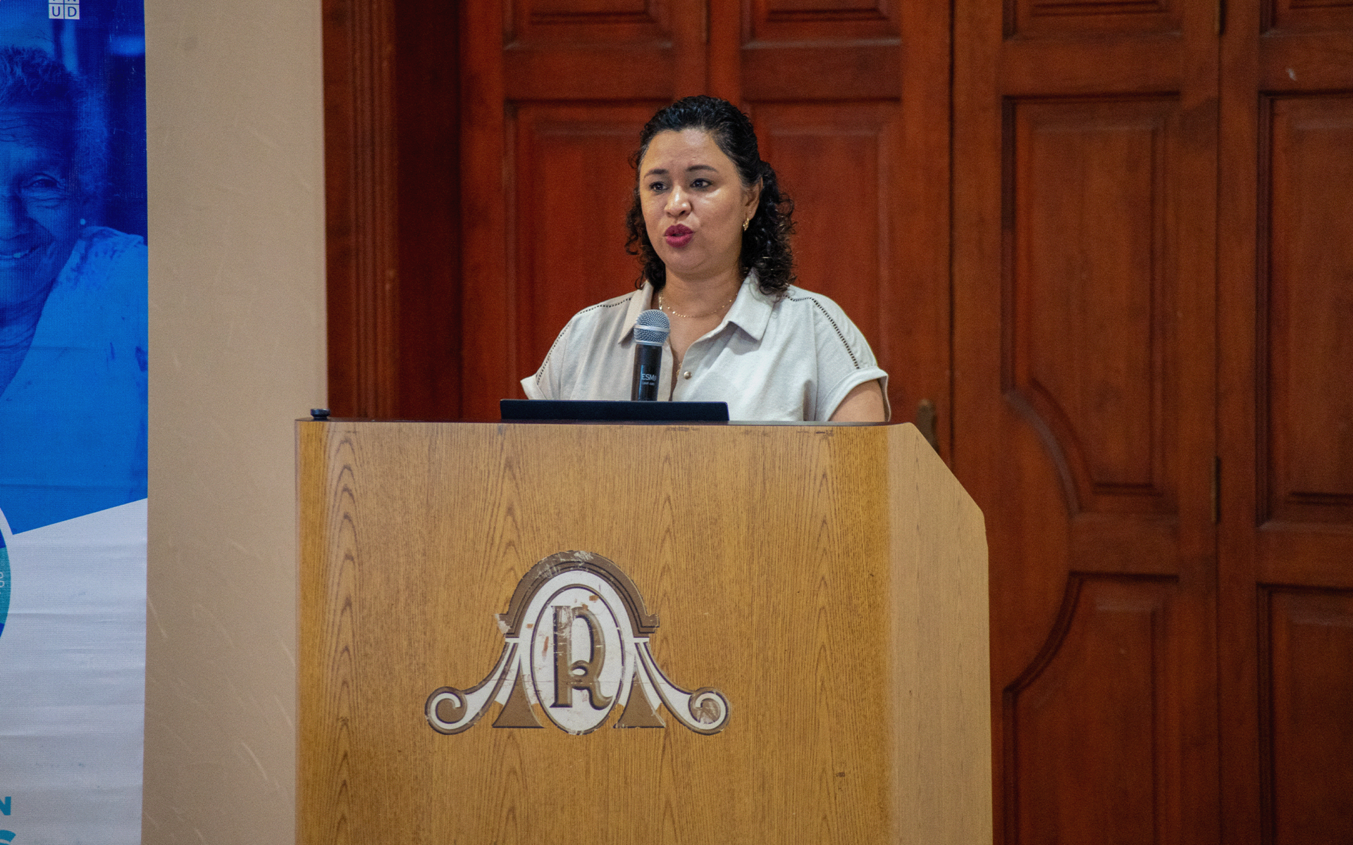 Woman delivering speech at wooden podium with crest in a paneled room.