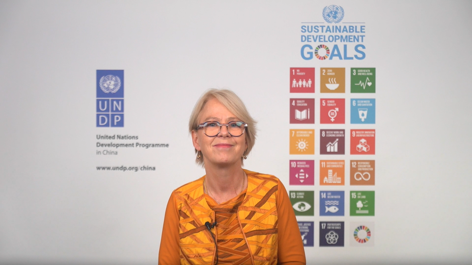 Person in orange jacket stands in front of a wall displaying Sustainable Development Goals icons.