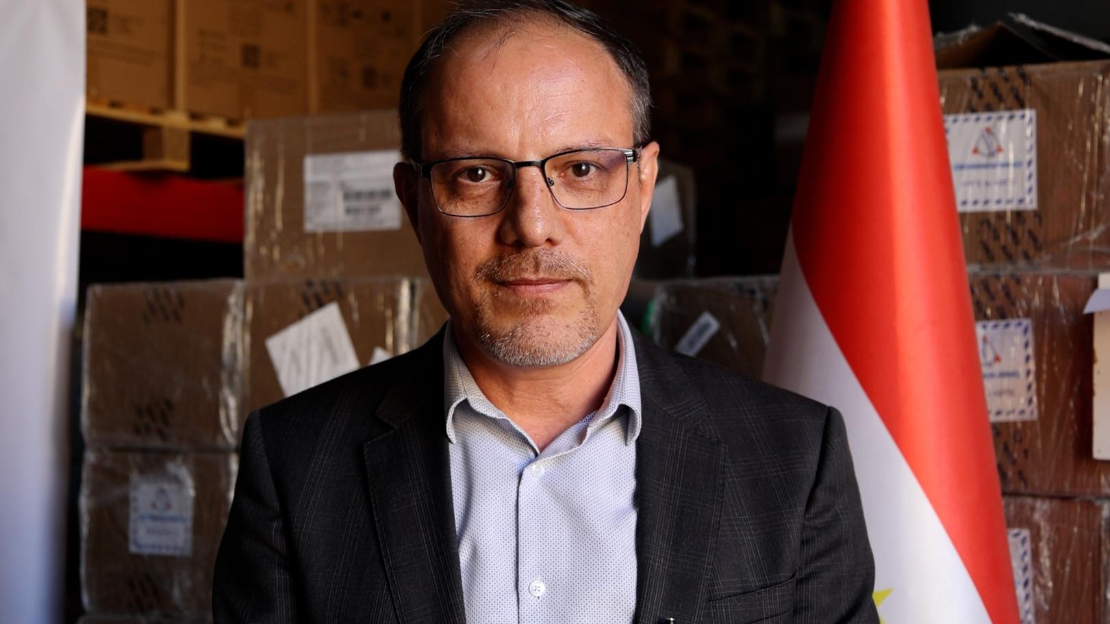 Man in a dark suit and light shirt stands in a storage area with boxes and a red flag.