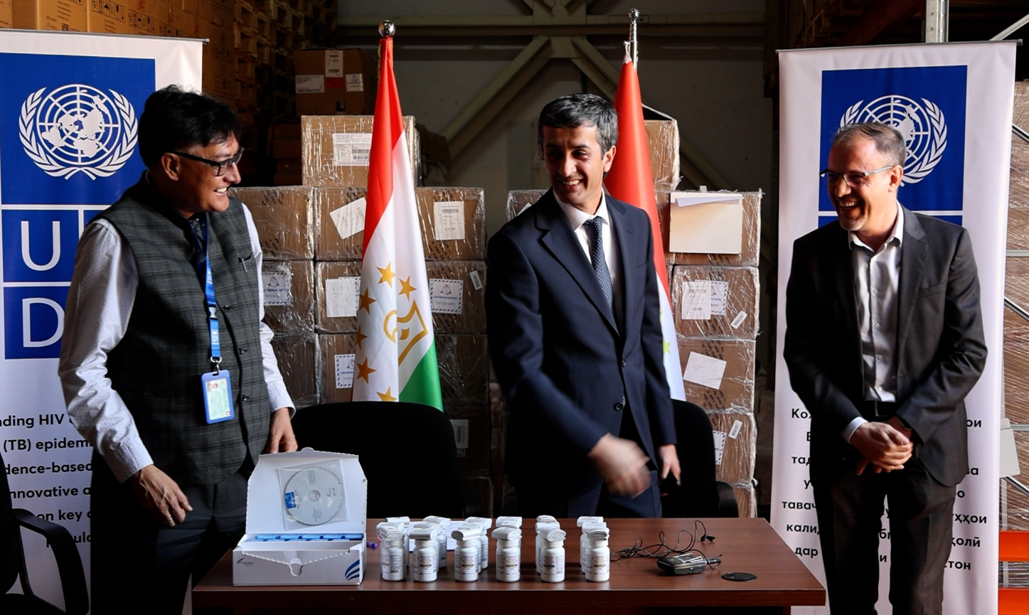 Photograph: Four men in suits stand around a table with cups; UN banners and flags in background.