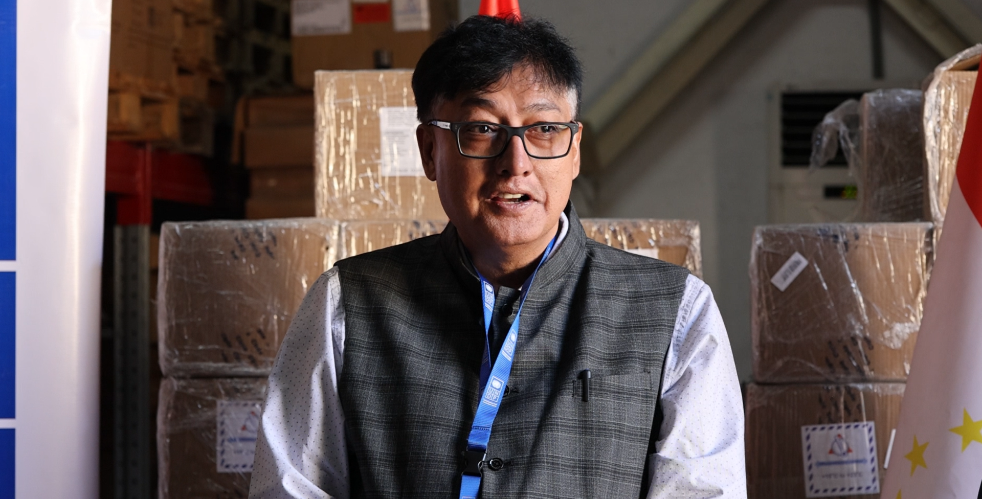 Photograph of a person in a gray vest with a blue lanyard in a warehouse with stacked boxes.