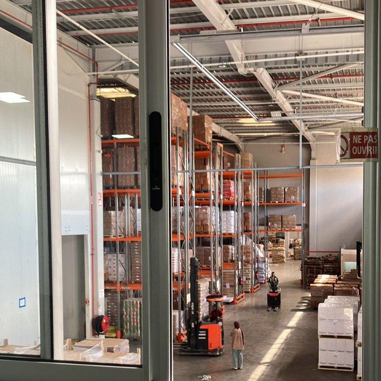 Warehouse interior with tall metal racks, pallets, and boxes; workers in orange vests.