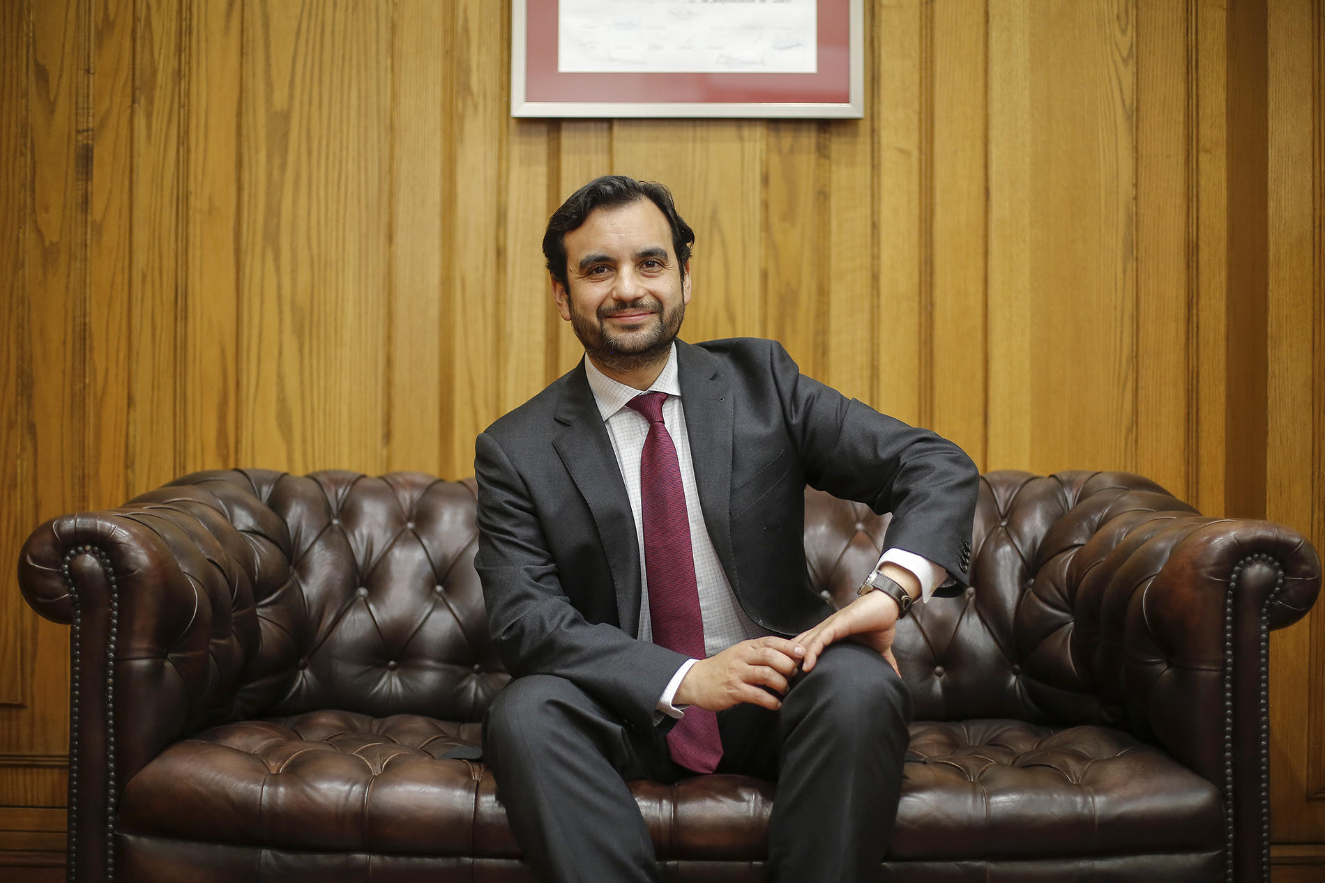 Man in a dark suit and red tie sits on a brown leather sofa in a wood-paneled office.