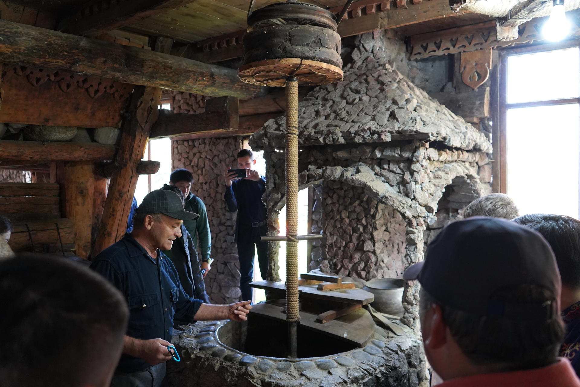 Photograph of people gathered around a large circular stone oven in a rustic log cabin kitchen.