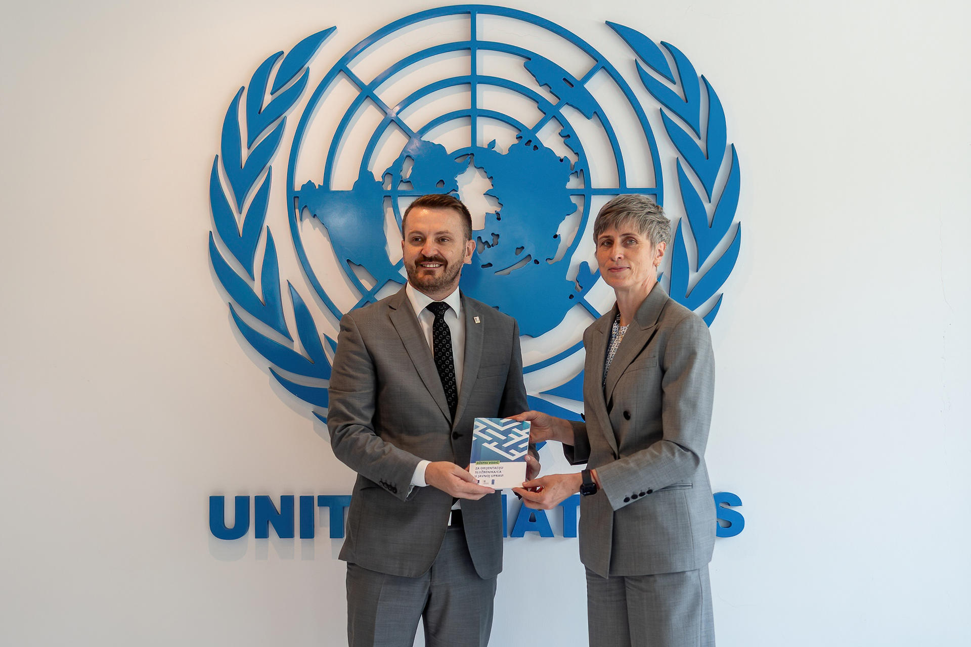 Two men in suits exchange a document in front of a United Nations emblem.
