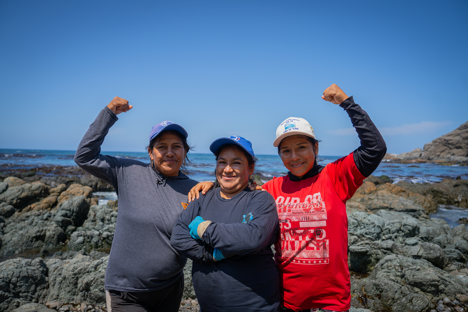 Three people on a rocky coastline with the blue sea behind, arms raised in celebration.