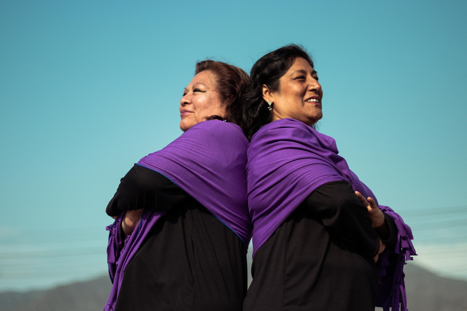 Two people in purple shawls standing back-to-back under a blue sky.