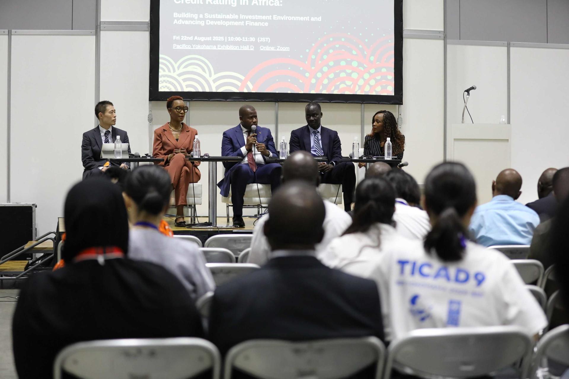 Panel discussion on stage with six speakers; audience seated in a conference room.