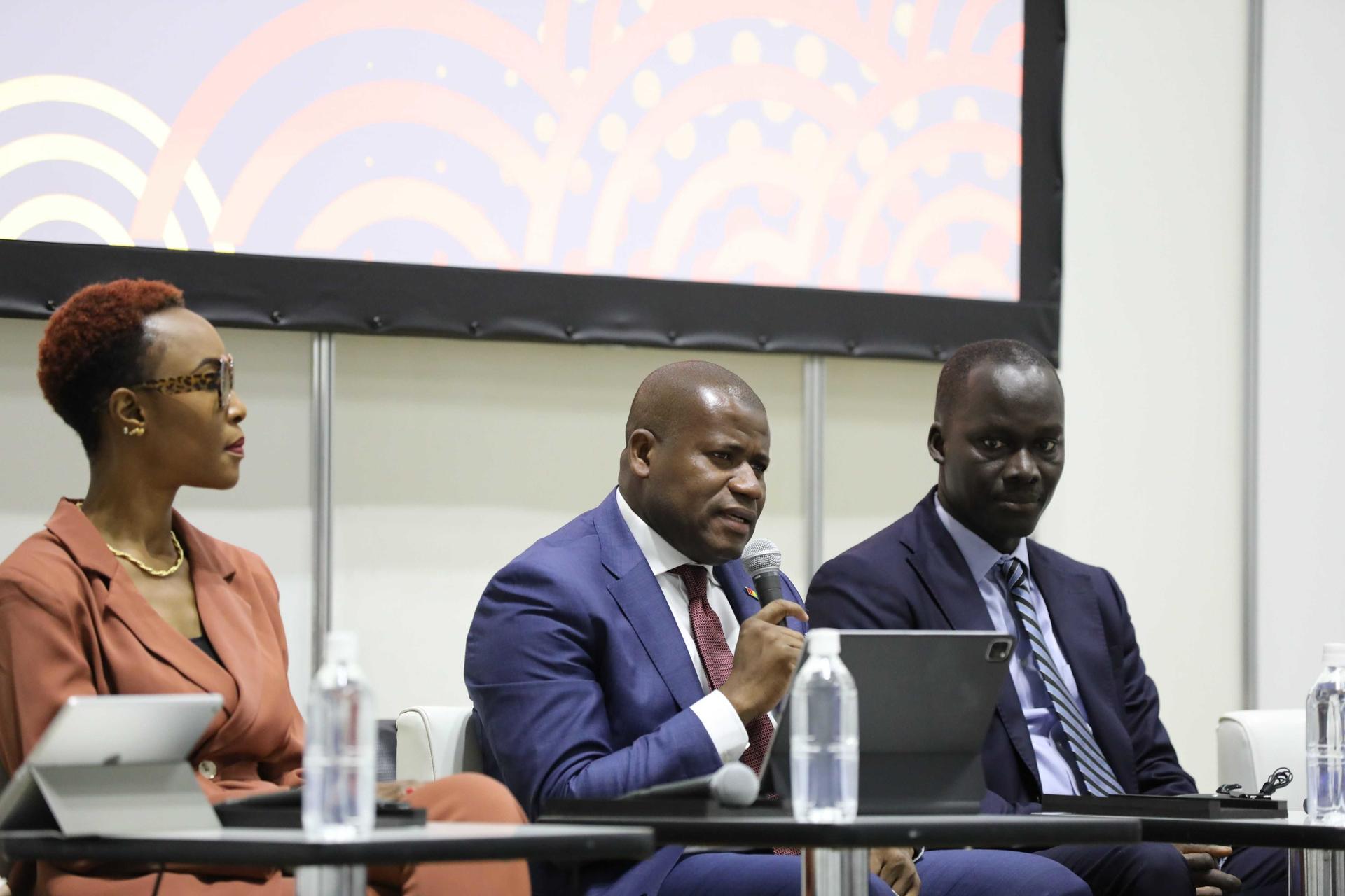 Three panelists in a conference room sit at a table with water bottles; screen in background.