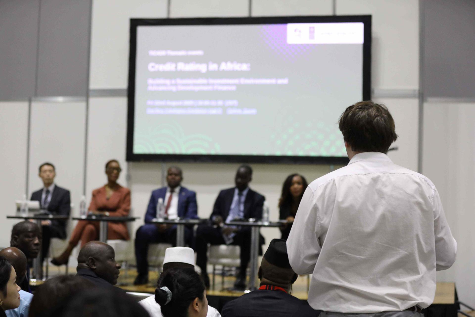Photograph shows panelists on stage; a man in the foreground addresses them.