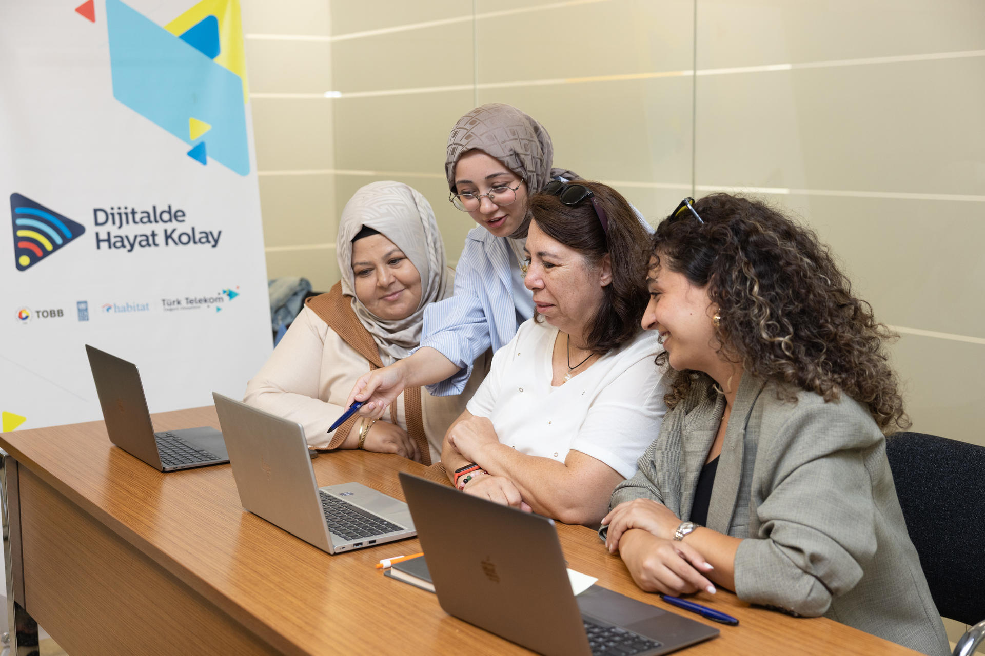 Group of five people at a training workshop using laptops; banner and glass wall in the background.