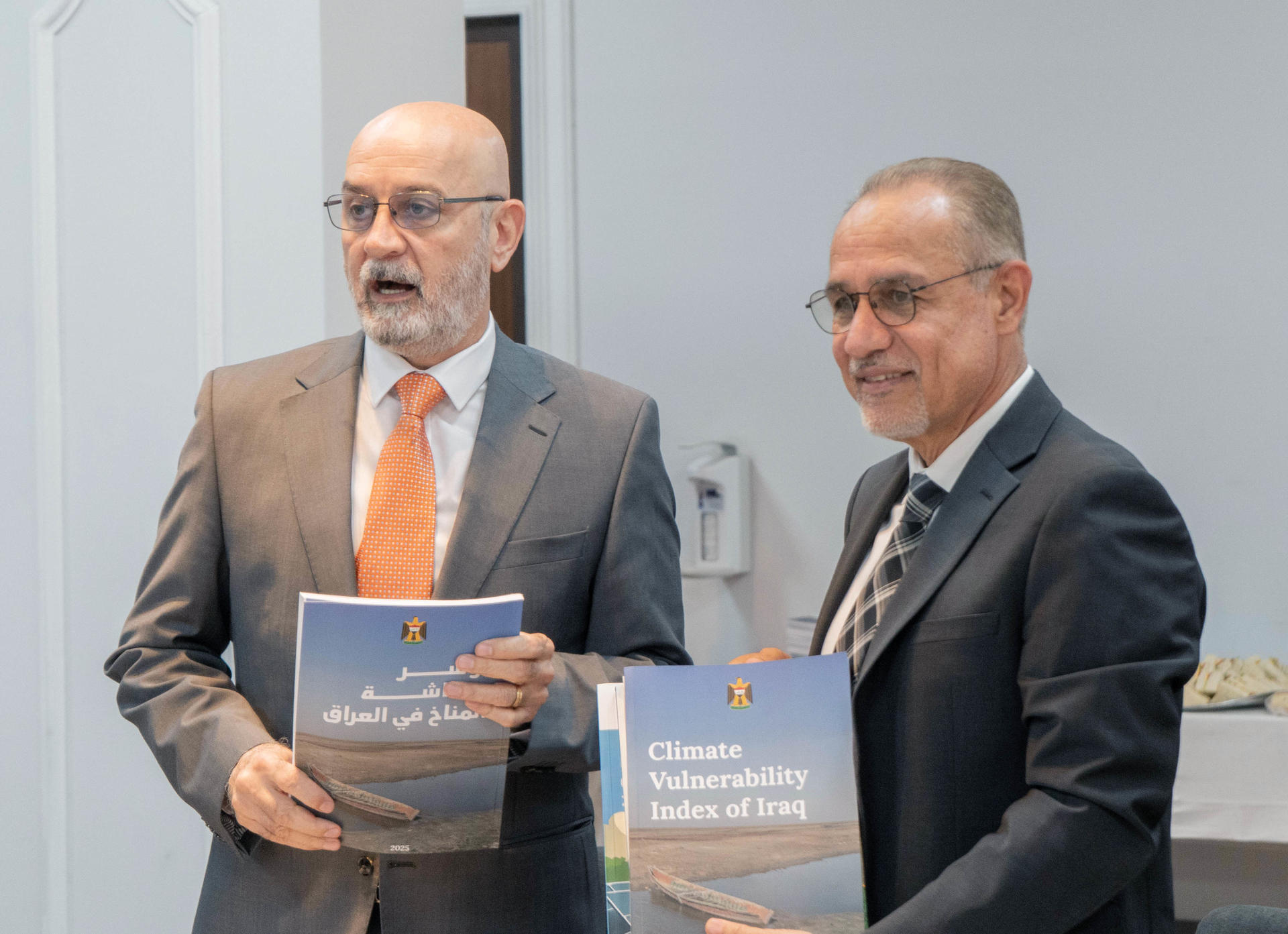 Two men in suits stand in a hallway, each holding a blue book.