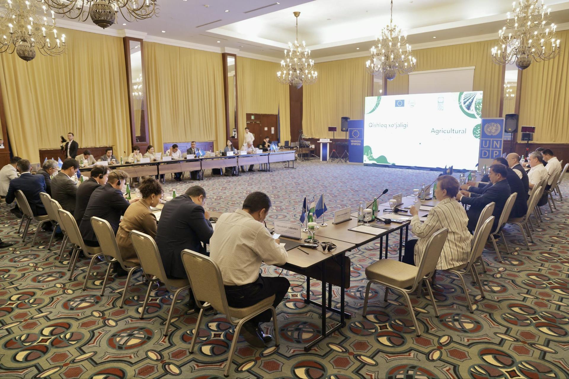 Large conference room with participants seated around U-shaped tables, projection screen at front.