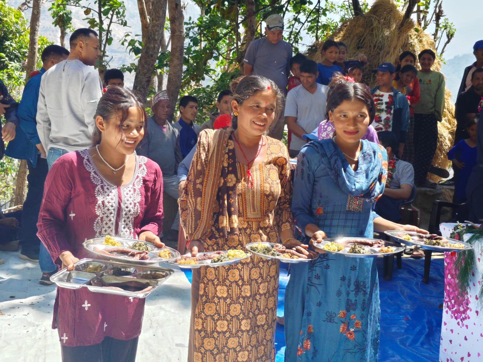 Three women in colorful traditional outfits carry plates at an outdoor festival, with crowd behind.