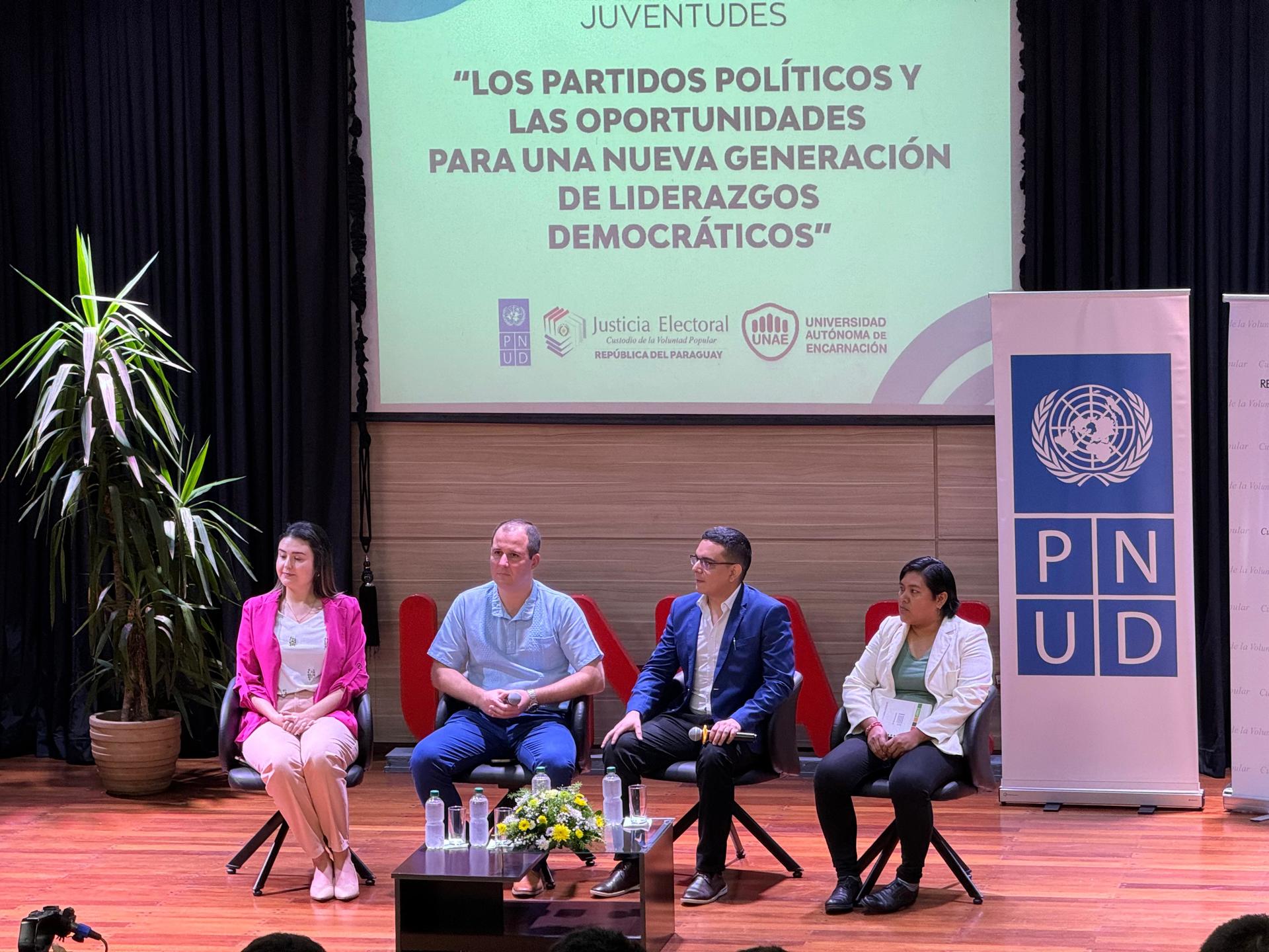 Five speakers seated on a stage during a panel discussion, with banners and a plant behind.