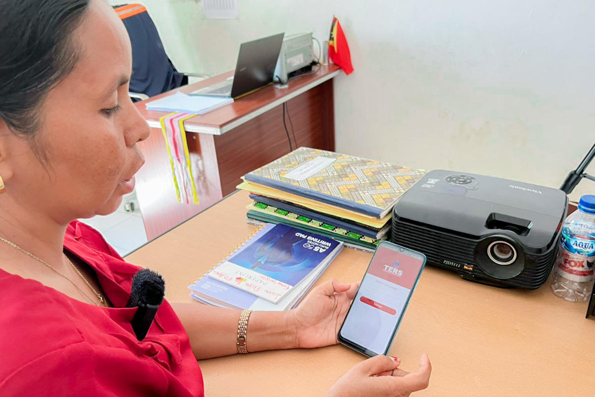 Person in red shirt at desk with laptop, printer, and papers, holding a smartphone showing a red-and-white screen.