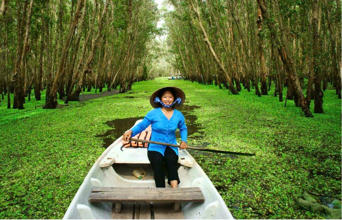 Person in blue shirt and wide-brim hat sits in a white canoe on a tree-lined canal.