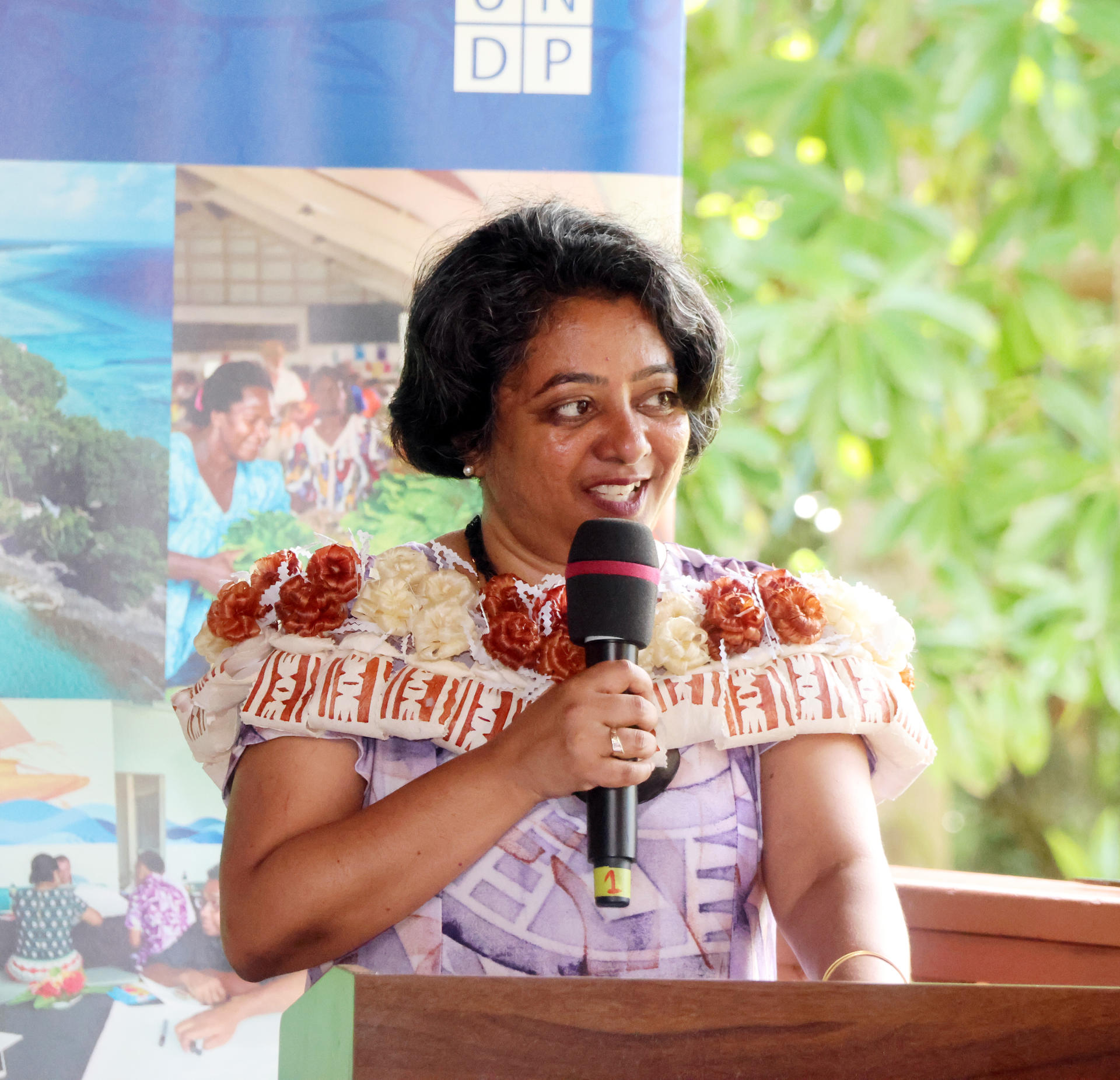 Speaker in traditional attire at a podium, outdoors, with banners in the background.