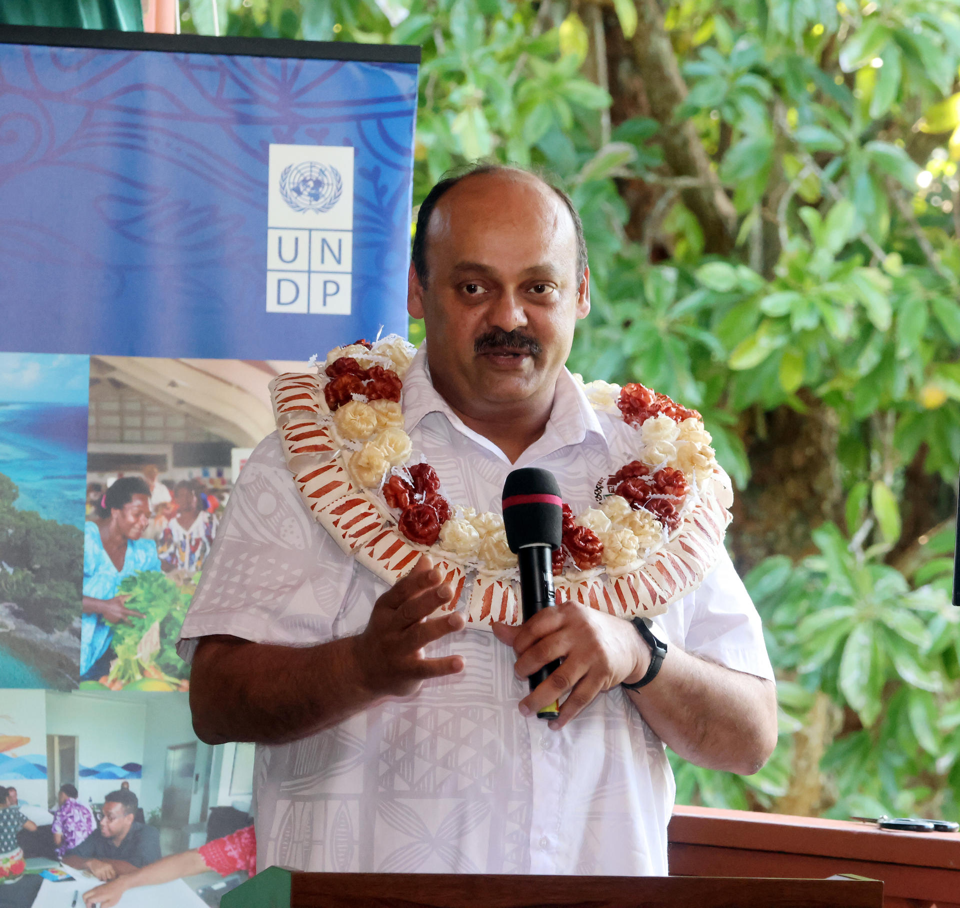 Man wearing a flower lei speaks at a podium with a UNDP banner in a leafy setting.