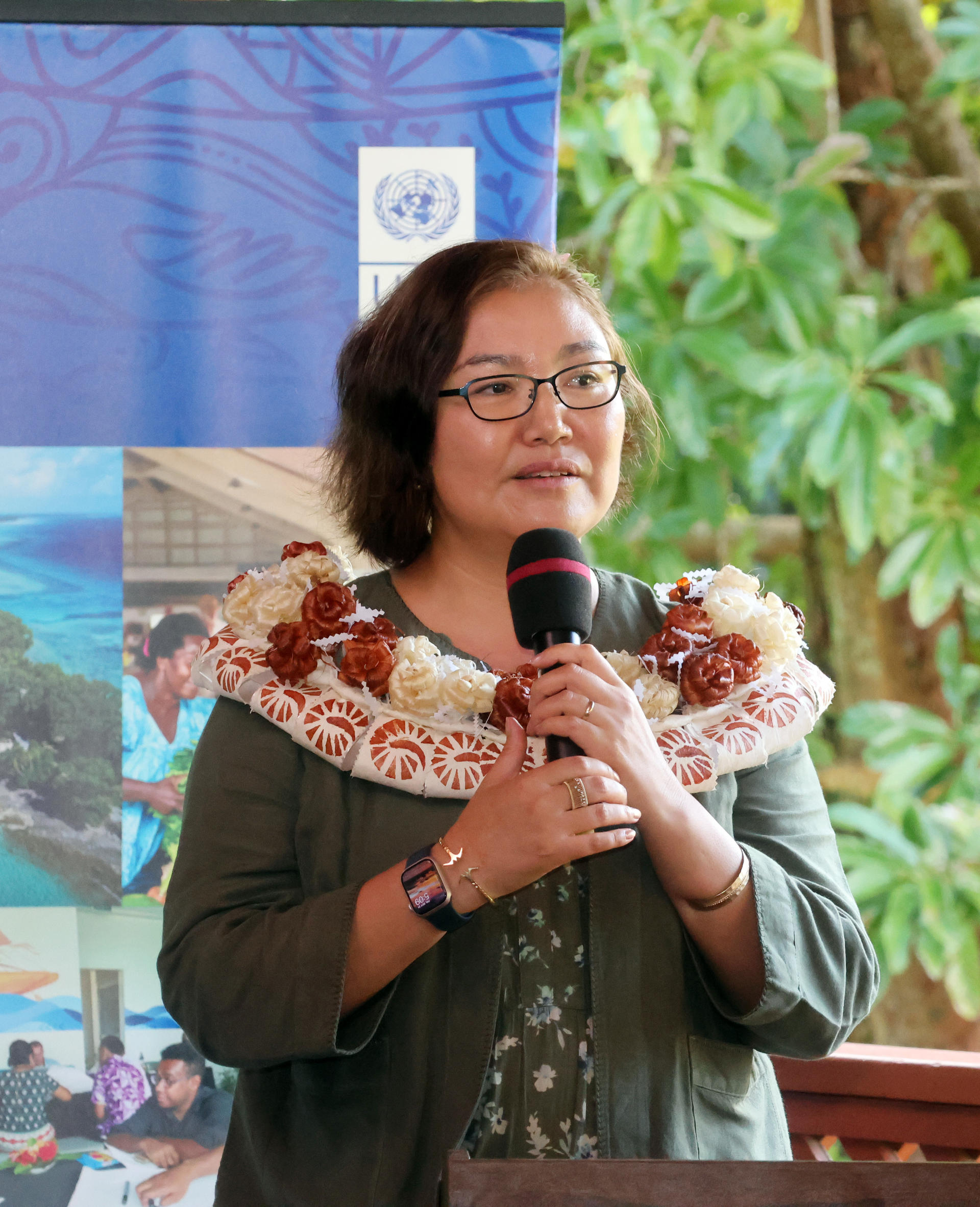 Speaker at a podium with microphone, wearing a floral shawl, in front of a blue banner.