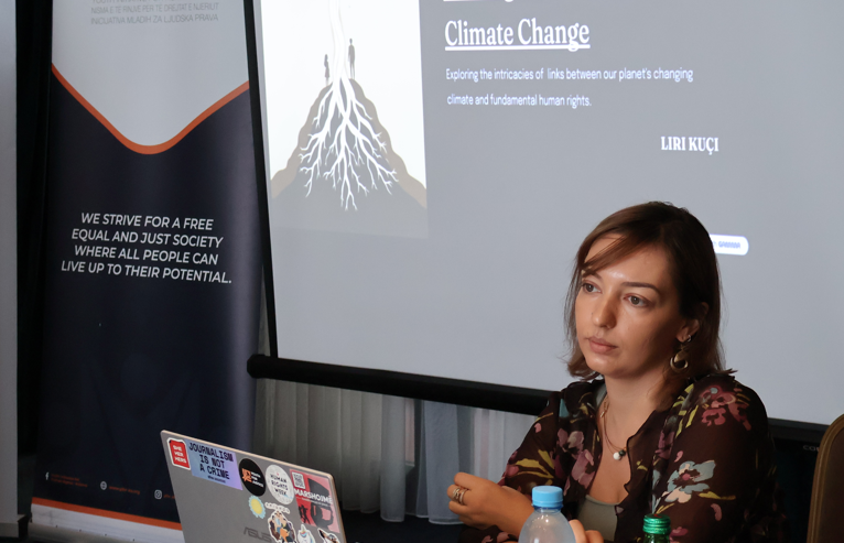 Presenter at a conference with microphone and laptop; screen displays Climate Change slide.