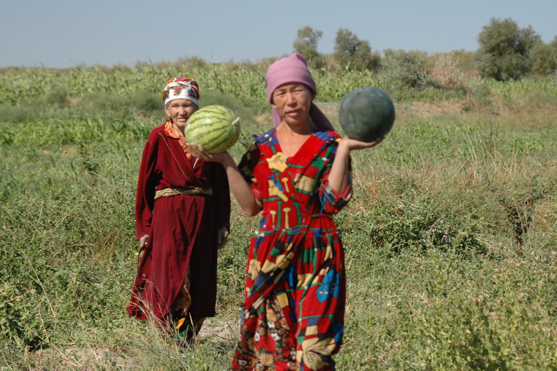 Two women in a rural field; one in a colorful dress holding a watermelon, the other in red robes.