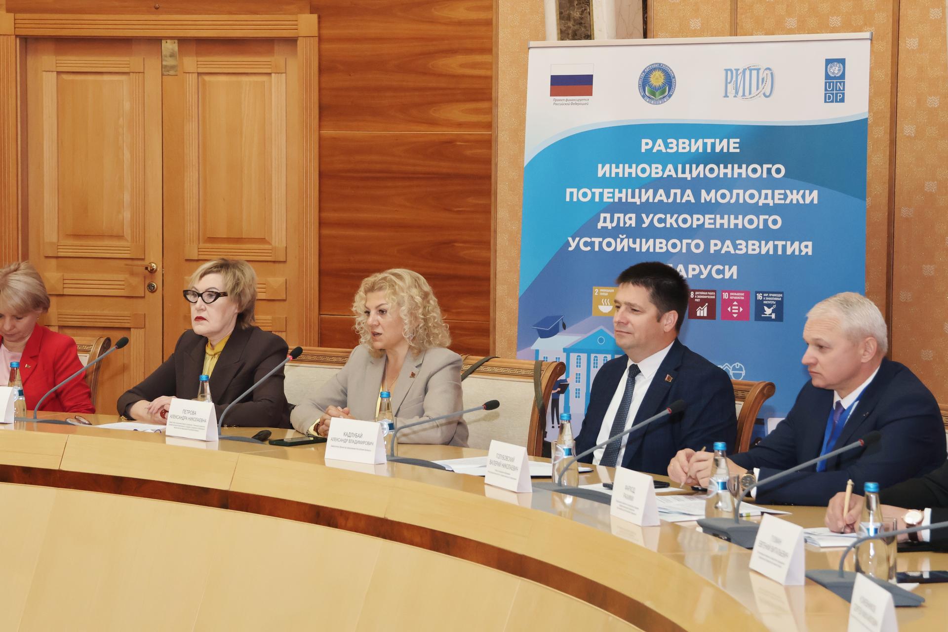 Six panelists at a round table with microphones; wood paneling and blue Cyrillic banner behind.