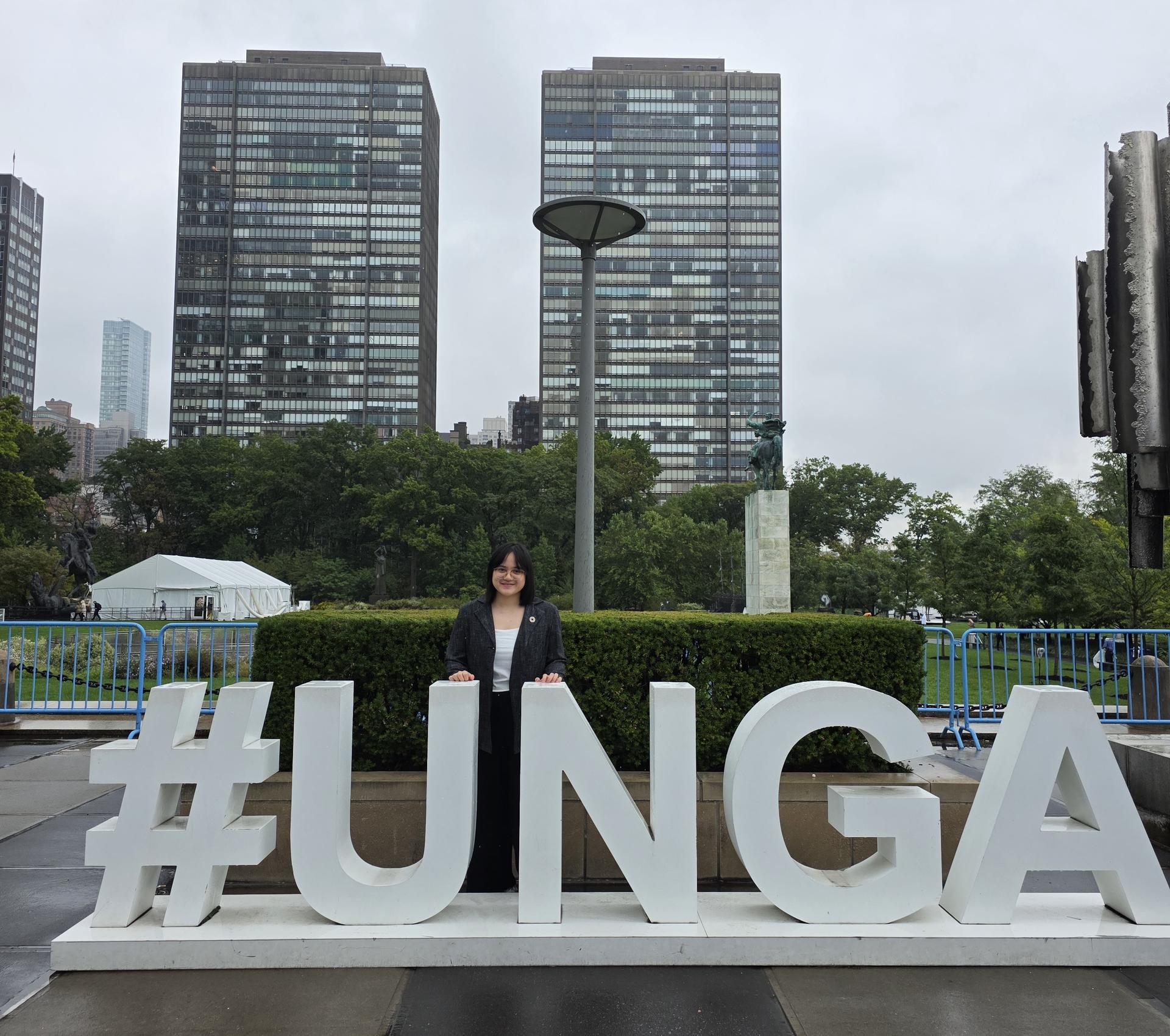Woman stands behind large white #UNGA letters on a plaza with hedges and tall buildings nearby.